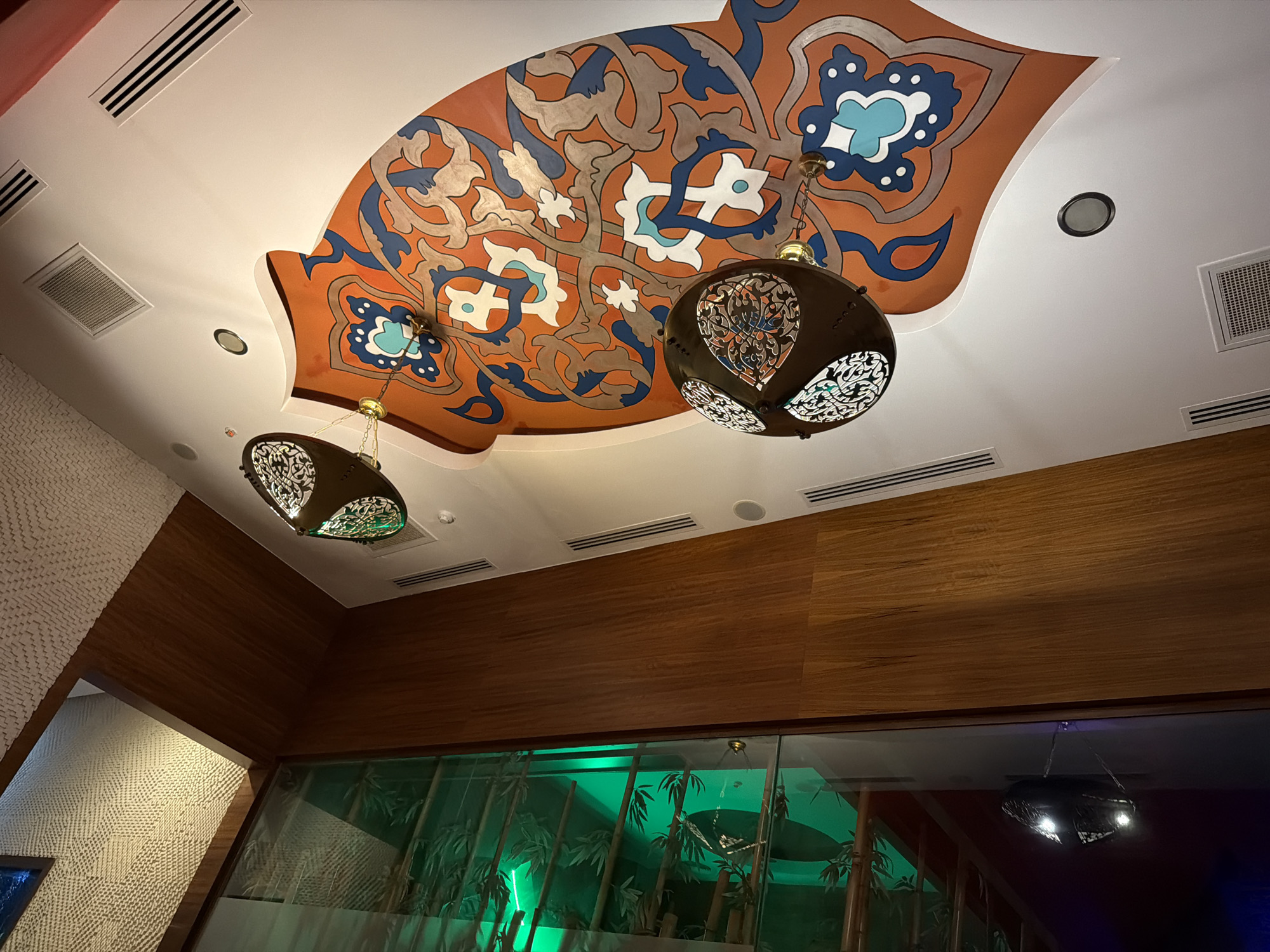 The image captures an interior scene with a decorative ceiling and lighting fixtures. The ceiling features a large, ornate mural in shades of orange, blue, and white, with intricate swirling patterns reminiscent of Islamic art. Two pendant lamps with detailed, dark metal shades hang from the ceiling, casting a warm glow. These lamps have an openwork design that allows light to filter through, creating interesting patterns.
In the background, a wall with a textured white finish is visible on the left, and a wooden-paneled wall runs horizontally across the upper portion of the image. Below the wooden paneling, a section of glass with a green tint reveals an indoor scene with bamboo-like plants, illuminated by bright green lights, suggesting a restaurant or lounge setting. Another dark pendant light fixture is visible reflecting in the glass in the lower right corner. The overall ambiance appears to be dimly lit and sophisticated, possibly in the evening. There is no visible text in the image. The scene is likely indoors, in a place designed for dining or relaxation. Given the context, this location could be Alanya, Turkey, a place known for its rich cultural influences reflected in its architecture and decor.