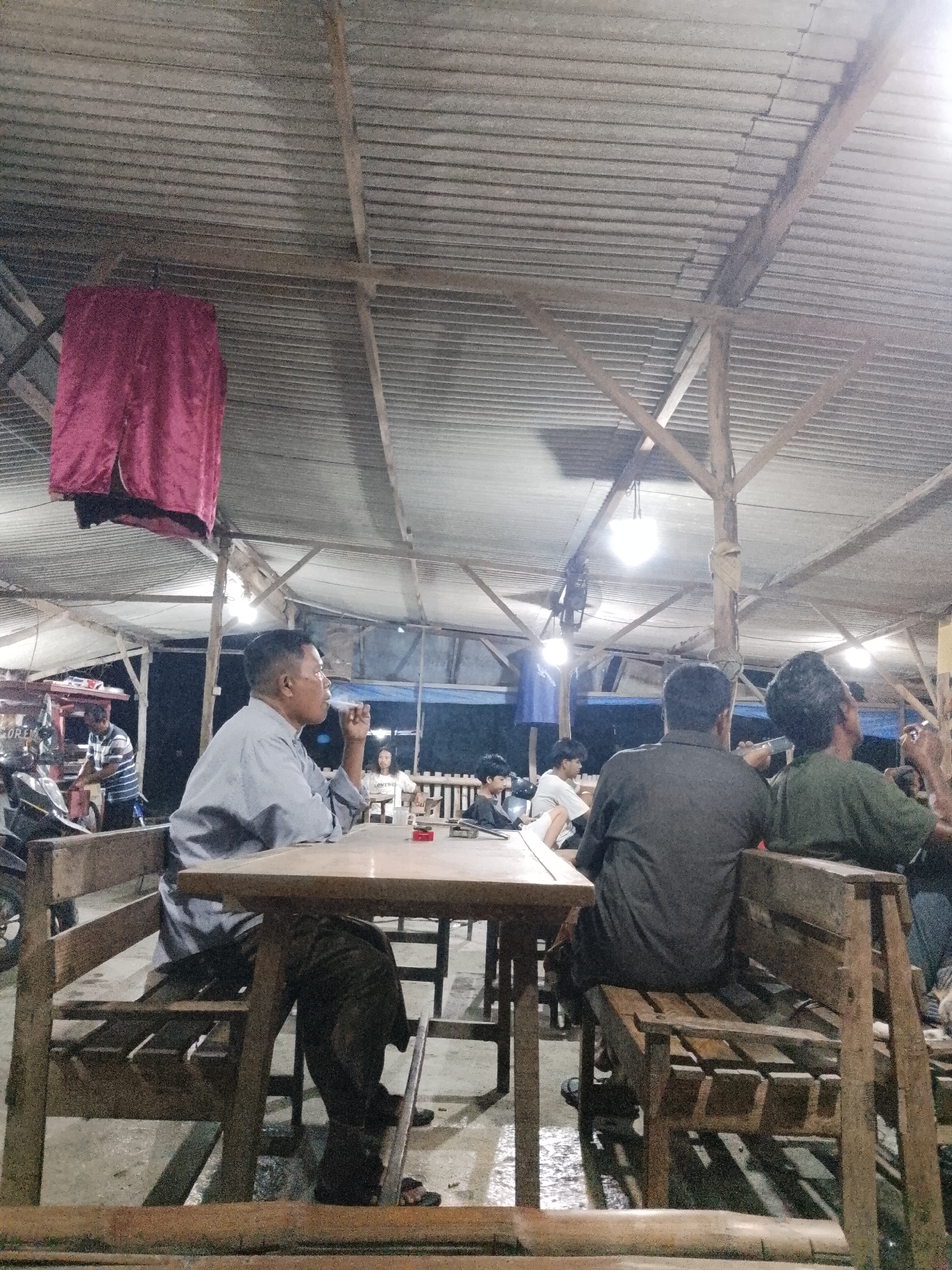 The image depicts an outdoor seating area of a casual eatery, likely a warung or similar establishment, in Sugihwaras, Indonesia. The scene is lit by artificial lights, suggesting it is evening or night. The structure has a corrugated metal roof supported by wooden beams and poles. A dark red fabric hangs from the ceiling on the left.
In the foreground, a man in a light blue shirt is seated at a wooden table, facing away from the camera and smoking. He appears to be in his 40s or 50s, with short, dark hair. His left elbow rests on the table, and he holds a cigarette to his mouth with his right hand. His attire is casual, with dark trousers.
Further back, several other individuals are visible. On the right side of the frame, two men are seated on a wooden bench, facing away from the camera. The man on the left wears a dark grey shirt, and the man on the right wears a greenish-brown shirt. They appear to be engaged in conversation or observing something.
In the middle ground, a person in a striped shirt is standing near a counter or cart, possibly preparing food or serving customers. A motorcycle is parked to the left of this person. Other patrons are seated at tables in the background, some appearing to be younger individuals. The ground appears to be concrete or a similar hard surface, with some puddles or wet patches visible, indicating recent rain or cleaning. The overall atmosphere suggests a relaxed, informal gathering place for local residents.