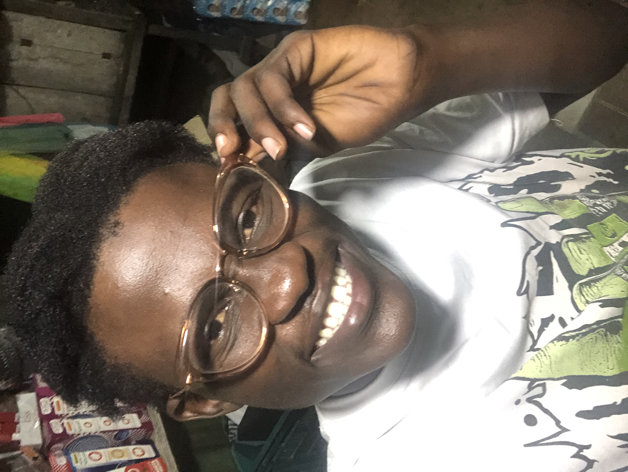 A close-up, slightly overhead selfie shows a dark-skinned person with short, dark, curly hair, wearing clear-rimmed glasses and a white t-shirt with a graphic print. The person is smiling broadly, revealing straight, white teeth, and is holding the right temple of their glasses with their right hand. The background is dimly lit and appears to be an indoor environment. On the left, shelves are visible, stocked with boxed goods. In the center background, behind the person's head, are colorful fabrics hanging, and to the right, a wooden structure is visible, suggesting a stall or shop. The overall impression is that the photo was taken in a marketplace or informal commercial setting in Ado, Nigeria.