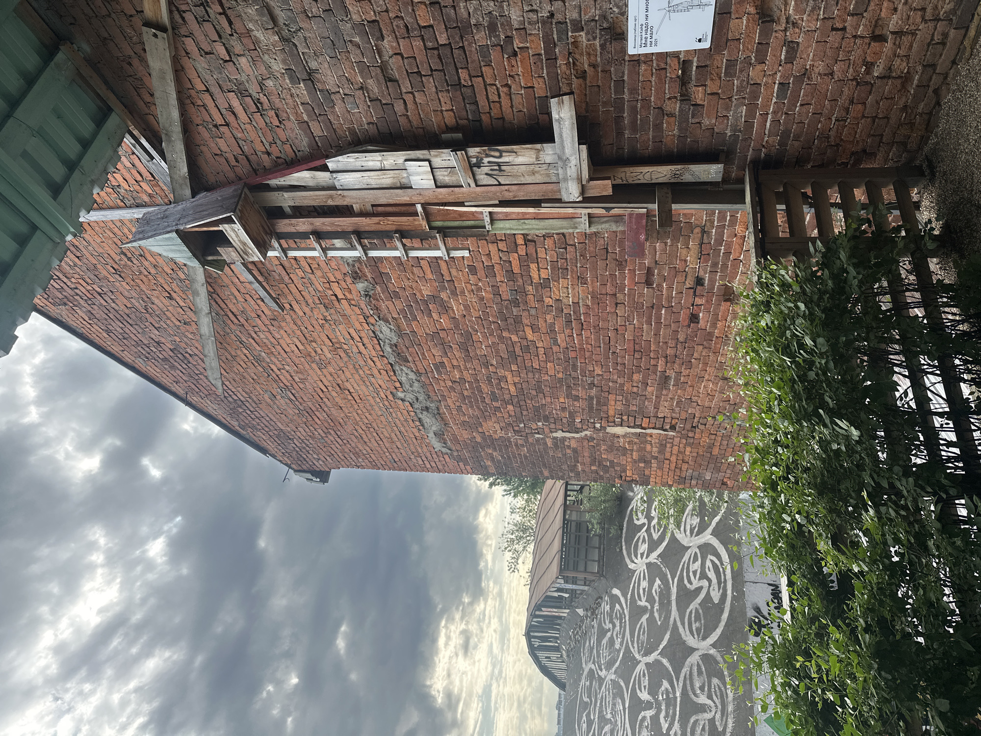 The image shows a brick archway in Moscow, Russia. The brickwork is weathered and aged, with visible cracks and repairs. Wooden planks and beams are affixed to the underside of the arch. A birdhouse is also attached to the arch. The archway opens onto a street or walkway. Graffiti art in the form of repeating circular patterns is on the street. The sky is overcast, with dark clouds. Green foliage is present to the right of the arch.