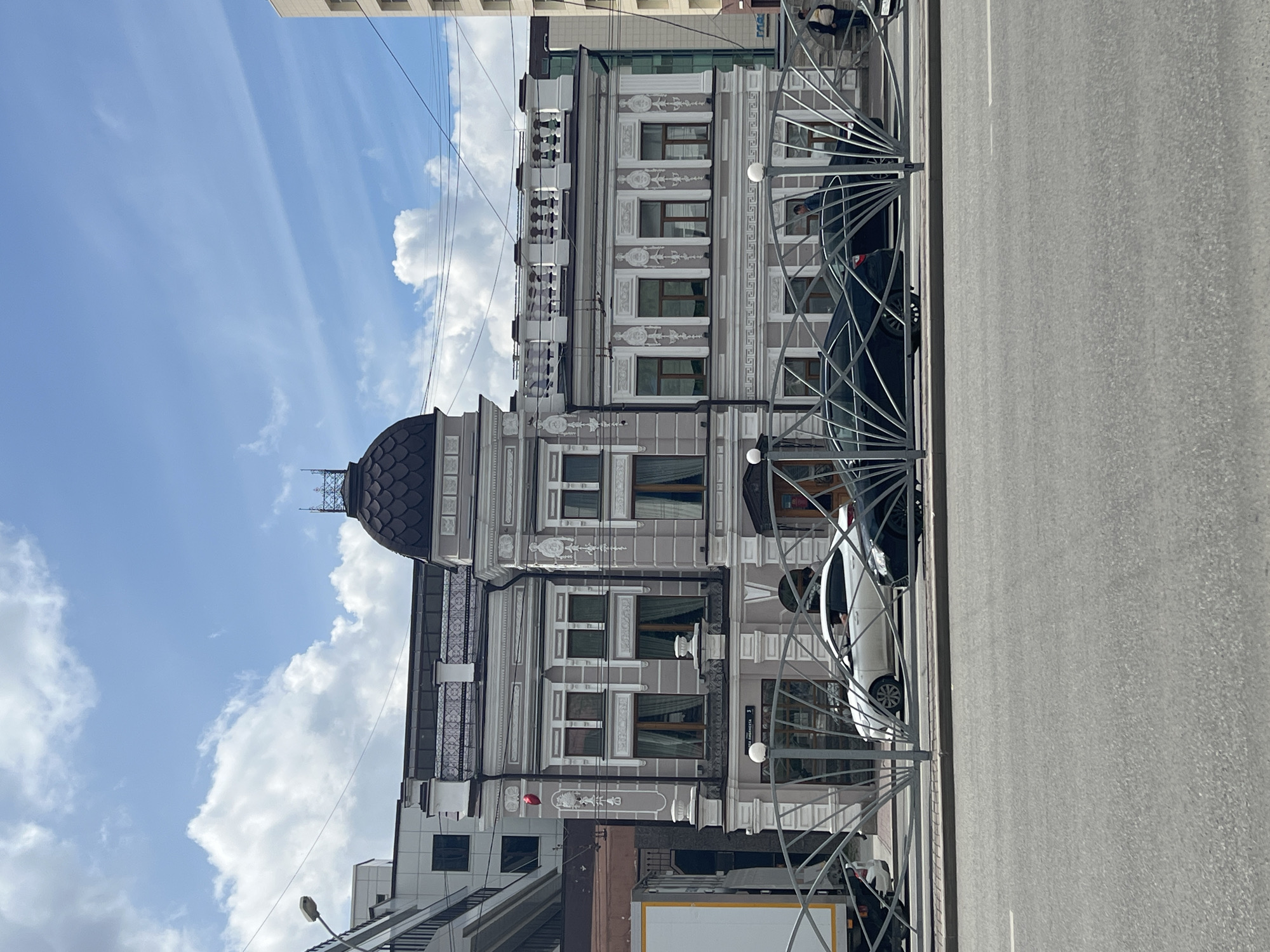This is a street-level, eye-level, medium shot photograph of a historic building in Yekaterinburg, Russia, on a bright, cloudy day. The building, a light grey structure with white ornate detailing, features a prominent dark dome with a decorative finial. The facade is characterized by numerous windows with white frames and light brown curtains, as well as decorative columns and balconies. A white railing runs along the foreground, partially obscuring a road where a white sedan and a dark sedan are parked. A white truck is visible on the left side of the frame. Several people are seen near the building entrance and on the sidewalk. The sky above is blue with scattered white clouds. The image appears to have been taken from across the street, looking towards the building.