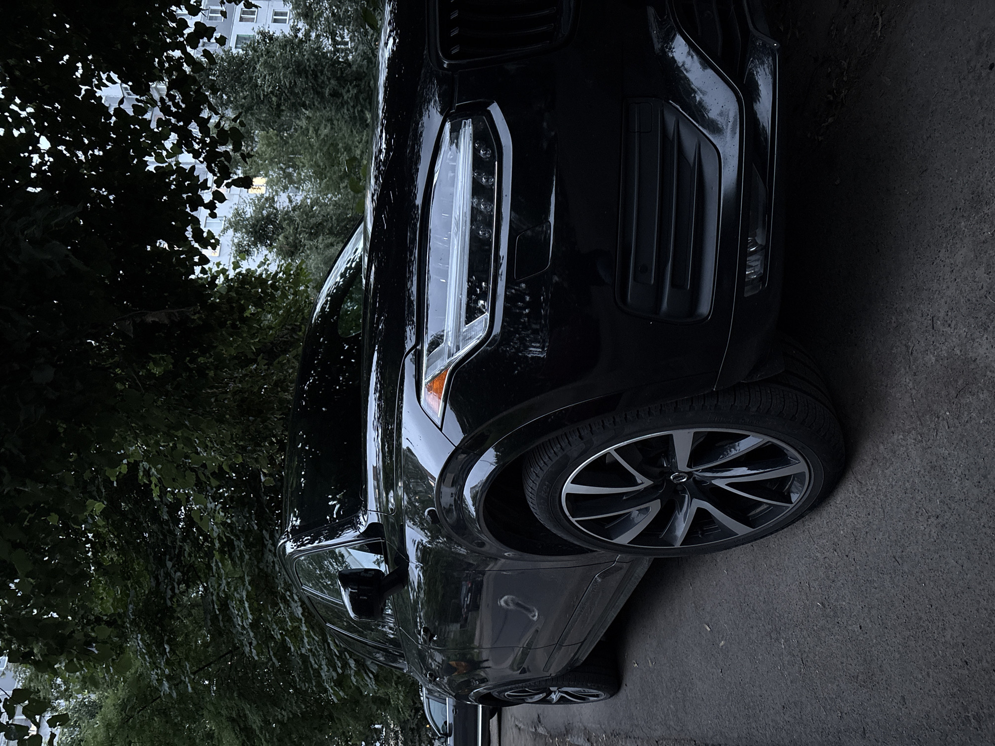 A black Volvo SUV is parked on a paved surface, possibly a street or parking lot, in Vitsebsk, Belarus. The car is positioned at an angle, with its front right side facing the camera. Lush green trees with dense foliage overhang the vehicle, casting shadows and creating a natural, outdoor setting. In the background, a building with multiple windows is visible through the leaves, suggesting an urban or suburban environment.
The image appears to have been taken during the day, though the overcast or shaded conditions contribute to a slightly dim and moody atmosphere. The car's exterior is wet, with water droplets visible on the hood and body, indicating recent rain or high humidity. The headlights of the SUV are illuminated, suggesting it might be dusk, dawn, or simply a very dark, cloudy day. The car features distinctive alloy wheels with a multi-spoke design. There is no visible text in the image. The scene conveys a sense of quiet stillness, with the focus primarily on the parked vehicle and its surrounding natural and man-made elements.
