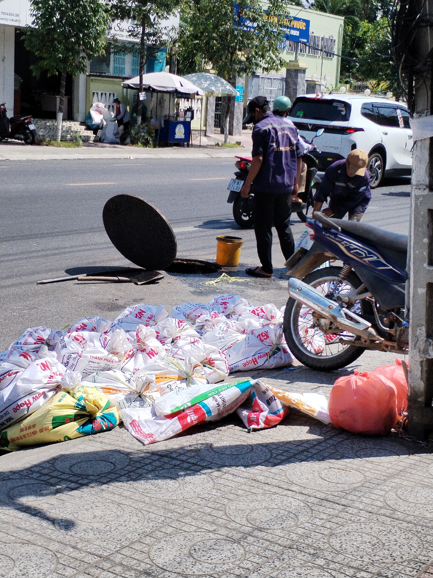 The image captures a scene on a street in Bac Nha Trang Ward, Vietnam, on a sunny day. In the foreground, a large pile of white, woven plastic bags, some appearing to contain agricultural products, is stacked on a paved sidewalk. Several of these bags are labeled with Vietnamese text and logos such as "New Hope." Among the bags, a yellow bucket and a metal shovel are visible, and a large, circular manhole cover is partially lifted, propped against the opening. A dark shadow stretches across the sidewalk from the bags and possibly an unseen object.
Further back, on the street, two men in dark uniforms are engaged in an activity near a parked red motorcycle. One man, wearing a black baseball cap, is standing, while the other, wearing a tan baseball cap, is bent over, appearing to interact with something on the ground or the motorcycle. A white SUV is parked behind them. To the left, a small white umbrella covers a stand or cart. Trees with green foliage line the street, and buildings with light-colored facades and blue shutters are visible in the background. The overall impression is a street scene in progress, possibly related to infrastructure work or delivery.