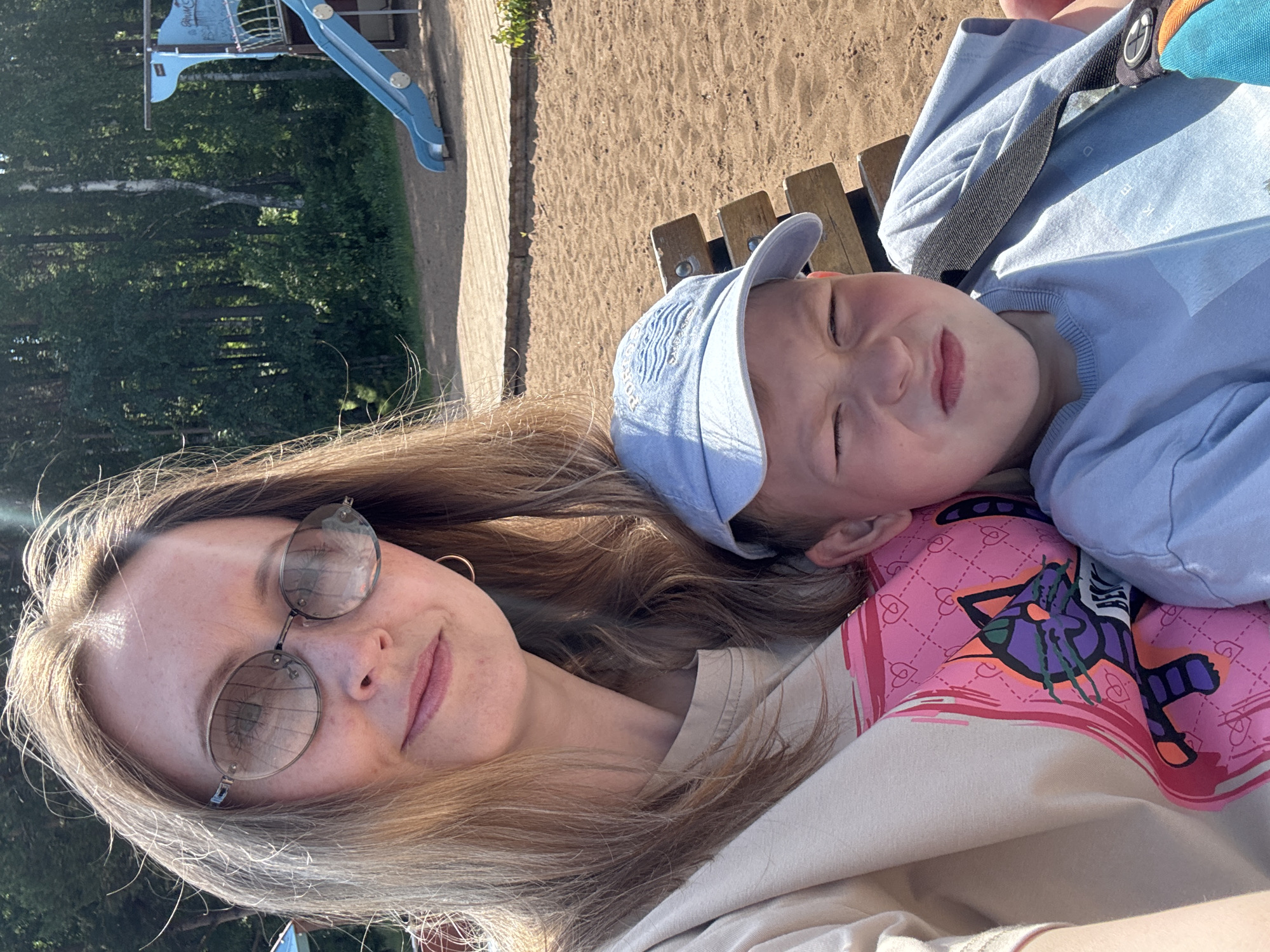 A woman and a young boy are seated on a wooden bench outdoors on a sunny day. The woman, on the left, has blonde hair and is wearing large, oval-shaped sunglasses and a light-colored t-shirt with a pink graphic of a cat. She is looking towards the camera. The boy, on the right, is wearing a light blue baseball cap and a light blue t-shirt. He is making a displeased facial expression, with his eyes squinted shut and his mouth in a pout. Behind them, a playground structure with a blue slide is visible, set against a backdrop of green trees. The ground is covered in sand and wooden planks. The scene appears to be a park or recreational area in the city of Chaikovsky, Russia.
