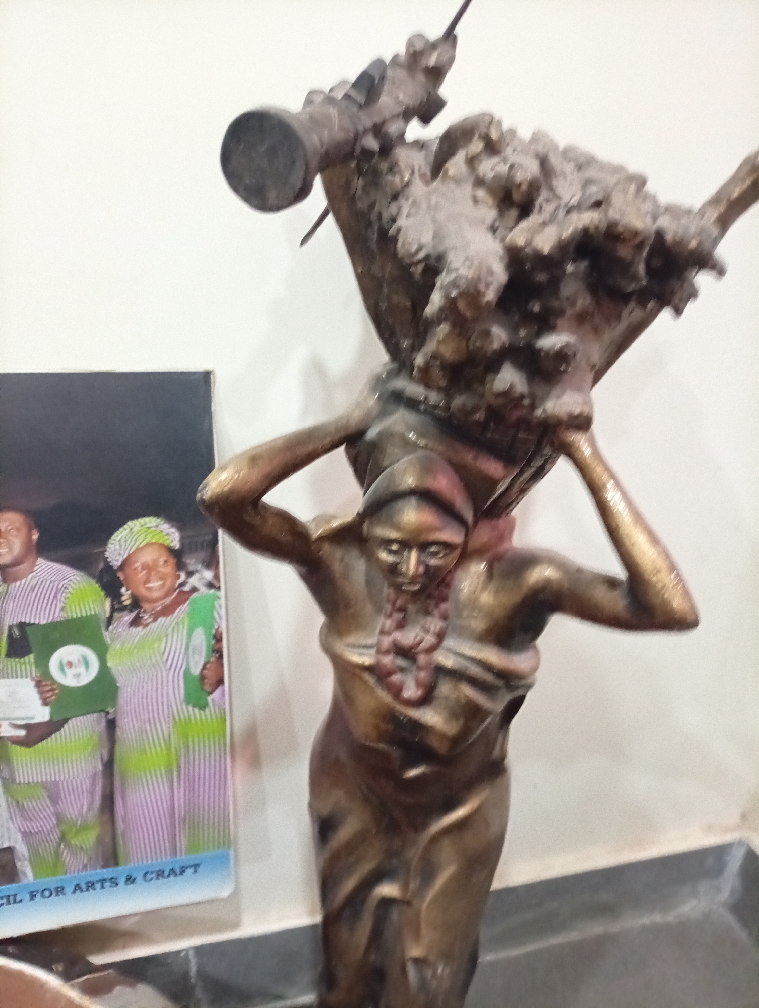 The image shows a bronze statue of a woman carrying a basket-like object on her head. The statue is in front of a book or framed picture that depicts two people, possibly dignitaries, holding green certificates. The text "CIL FOR ARTS & CRAFT" is visible at the bottom of the book. The location in the city of Dadi Riba, Nigeria is not readily identifiable from the image.