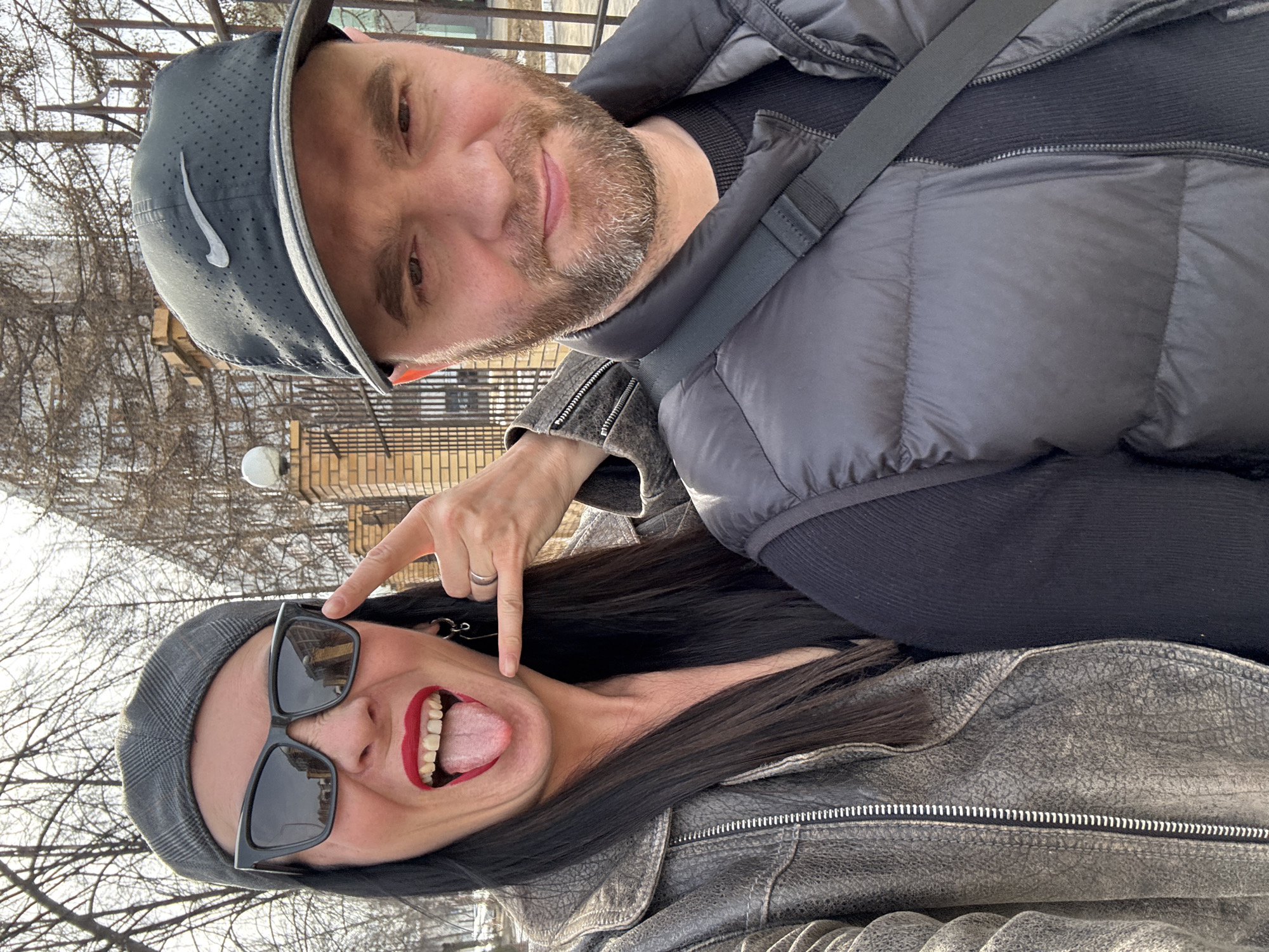 Two individuals are captured in an outdoor selfie. The person on the top, likely male, wears a gray Nike baseball cap, a black padded vest, and a black sweater. He has a beard and mustache. The person on the bottom, likely female, is wearing dark sunglasses, a gray cap, a gray leather jacket, and has dark hair. Her mouth is open, exposing her teeth and tongue, and she is using a finger to point at her face. The background shows trees without leaves, a brick fence, a lamppost, and buildings. Based on visual clues (architecture, streetlights) this photo appears to be taken in an urban environment in Chaikovsky, Russia.