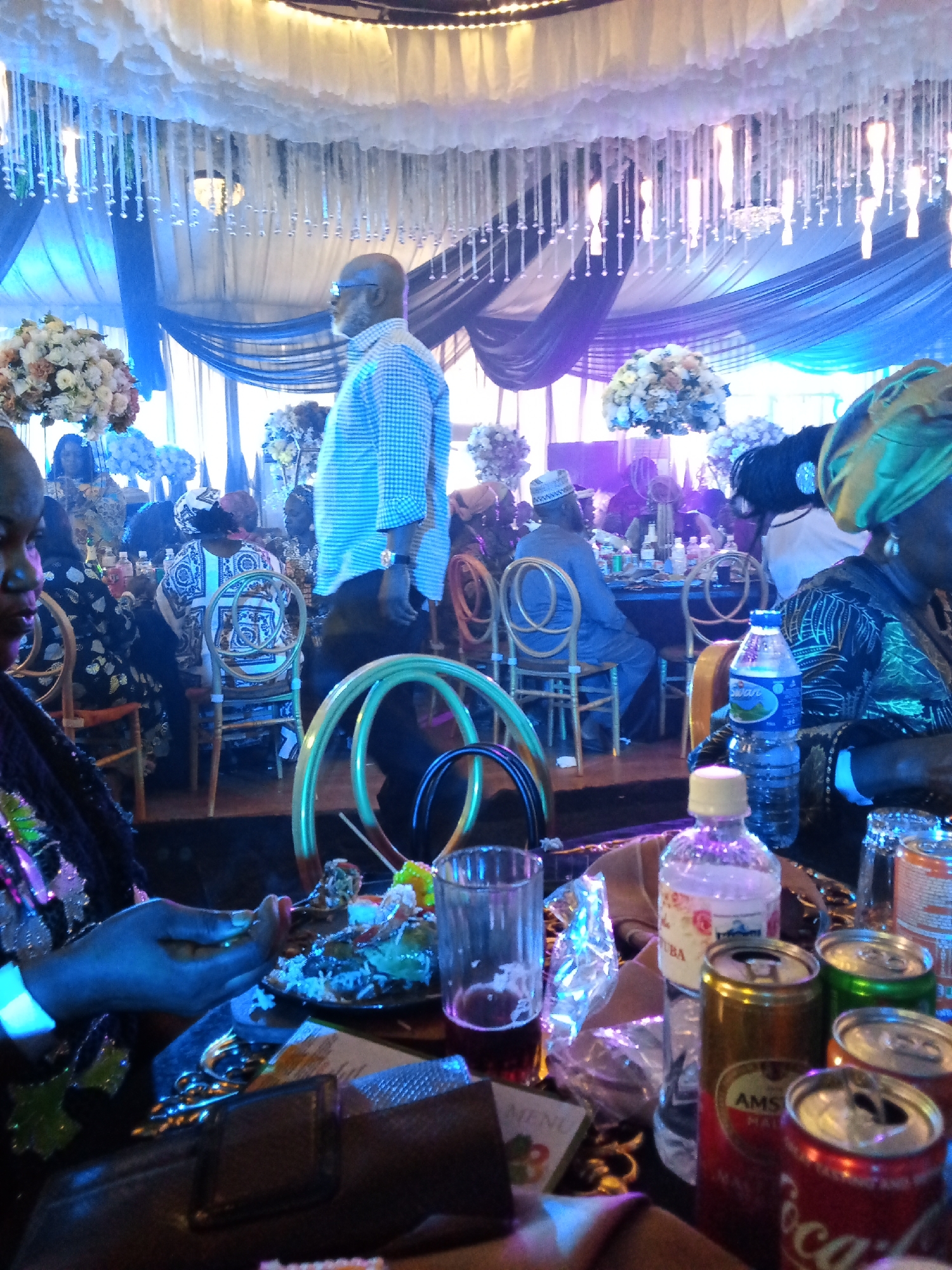 The image depicts a lively gathering, possibly a wedding reception or a celebration, taking place indoors under a decorated ceiling. The environment is richly adorned with white fabric clouds and crystal-like hanging ornaments, illuminated by ambient blue and white lights, creating a festive atmosphere. Several individuals are visible, some seated at tables adorned with floral arrangements, while others are standing or in motion.
In the foreground, a woman with dark skin is seen from the chest up, wearing a black garment with colorful, possibly sequined, floral embellishments. Her left hand is extended, holding a fork, and she appears to be eating from a plate of food, which is partially visible. Next to her, on the table, are several beverage cans and a glass of dark liquid, along with a bottle of water.
Behind her, a man in a blue and white checkered shirt is standing and looking to his right. He has a beard and is wearing glasses. Further into the background, more people are seated at tables, some of whom are dressed in traditional attire. One woman wears a vibrant green headscarf and a patterned dress. Another man is seated in a chair, wearing a cap and a grey traditional outfit. The tables are covered with dark tablecloths, and some are set with dishes and drinks. The overall impression is one of a well-attended event with a celebratory mood. The scene appears to be consistent with a Nigerian cultural event, possibly in a city like Barden Yana, known for its vibrant social gatherings.