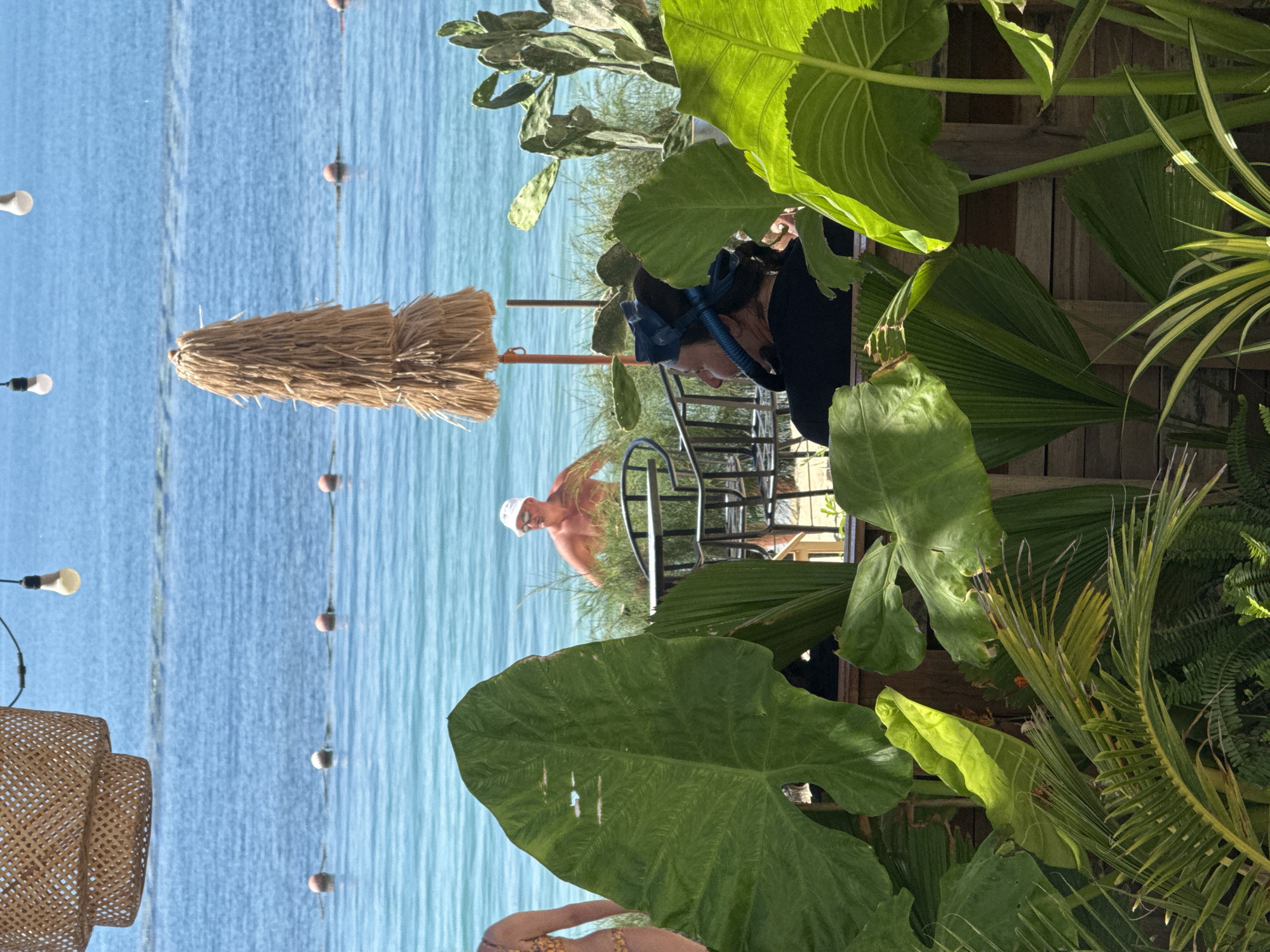 The image depicts a beachside scene in Nha Trang, Vietnam. In the foreground, lush green tropical foliage partially obscures the view. Behind the plants, a woman wearing a snorkeling mask and snorkel is seated, facing right. Further in the background, a man in a white bucket hat is standing in the shallow ocean water. The sea is calm and blue, with a line of buoys marking an area offshore. A thatched umbrella and a woven light fixture with two bulbs are visible. The overall ambiance suggests a relaxed tropical resort setting.