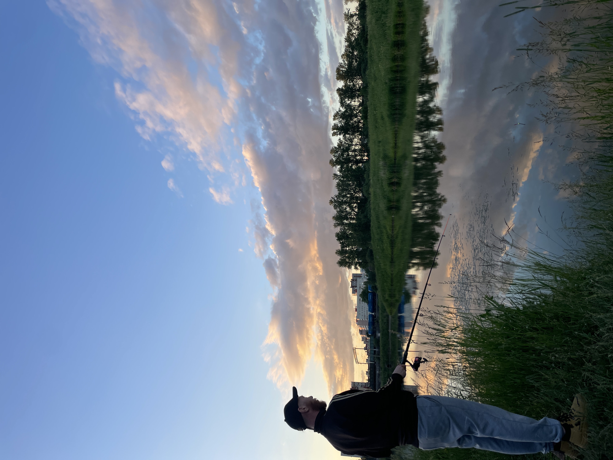 A man stands on the grassy bank of a river in Saint Petersburg, Russia, fishing as the sun sets. He is wearing a black jacket with stripes on the sleeves, light-colored pants, a black baseball cap, and tan hiking boots. He holds a fishing rod, casting his line into the water. The river reflects the soft colors of the sky, which is a mix of blue, pink, and orange clouds. In the background, across the river, is a line of trees and a distant cityscape with buildings and a train on tracks. The grass on the bank is tall and green, with some dried stalks mixed in. The water is calm, with small ripples disturbing the reflection of the sky and trees.