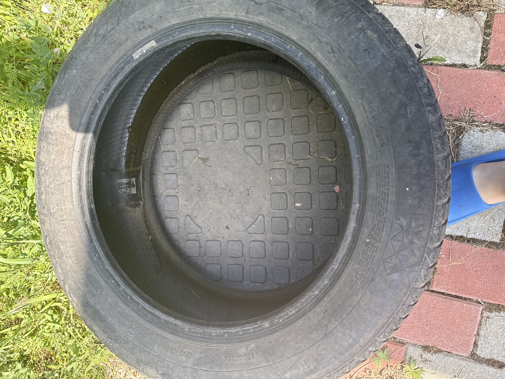 This media file features a top-down view of a car tire lying flat on the ground. The tire is black and appears to be used. Inside the tire, a metallic cover with a grid pattern of squares is visible. The tire is surrounded by elements of an outdoor setting, including green grass and red and gray paving stones. A small section of a blue flip-flop is visible in the lower right corner of the frame, suggesting a person's presence nearby. The lighting suggests it is daytime, likely outdoors in fair weather. Visible text on the tire includes "CONTINENTAL", "MADE IN GERMANY", "205/60 R16 96T", "MAX LOAD 710 KG (1565 LB)", and safety warnings.