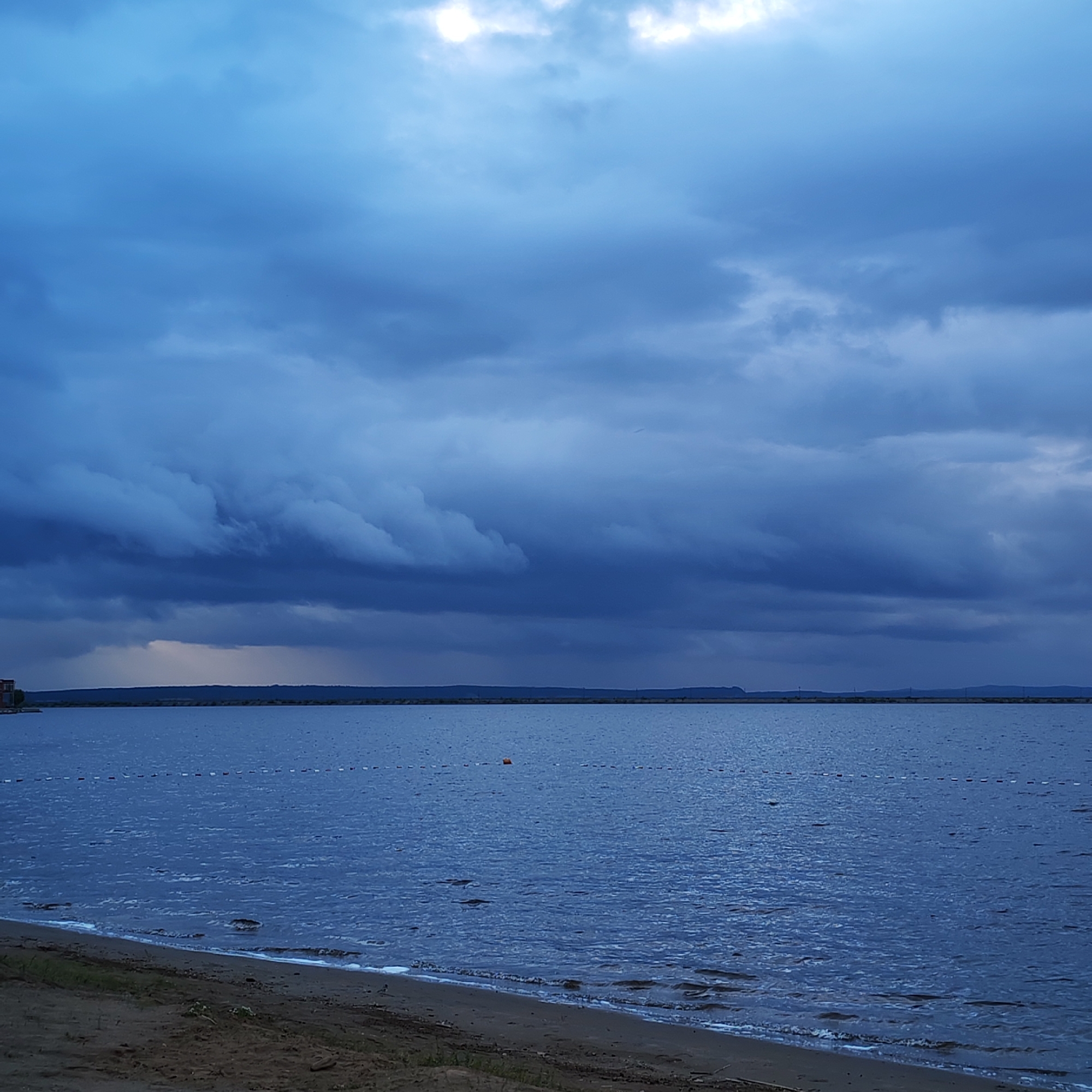 The image captures a vast, overcast landscape over a large body of water, situated in the city of Chaikovsky, Russia. The sky, occupying the upper two-thirds of the frame, is filled with heavy, dark blue-grey clouds. Lighter areas appear in the upper-center, suggesting obscured sunlight, while a visible curtain of precipitation descends from the clouds on the distant left, creating a hazy, lighter tone above the horizon. Across the mid-ground, a thin, dark line of land or distant shore stretches horizontally, with part of a reddish-brown building visible on the far left of this distant bank. The expansive body of water, consistent with a large reservoir like the Votkinsk Reservoir, is dark blue-grey and shows a choppy surface with ripples, indicating wind activity. A demarcation line consisting of small white and red buoys extends across the water in the mid-distance, with a single orange buoy positioned near the center of this line. The foreground features a dark, sandy beach sloping gently into the water, where small waves break, forming thin lines of white foam. Sparse green vegetation is discernible along the sandy edge on the far left. No individuals or active subjects are present within the scene.