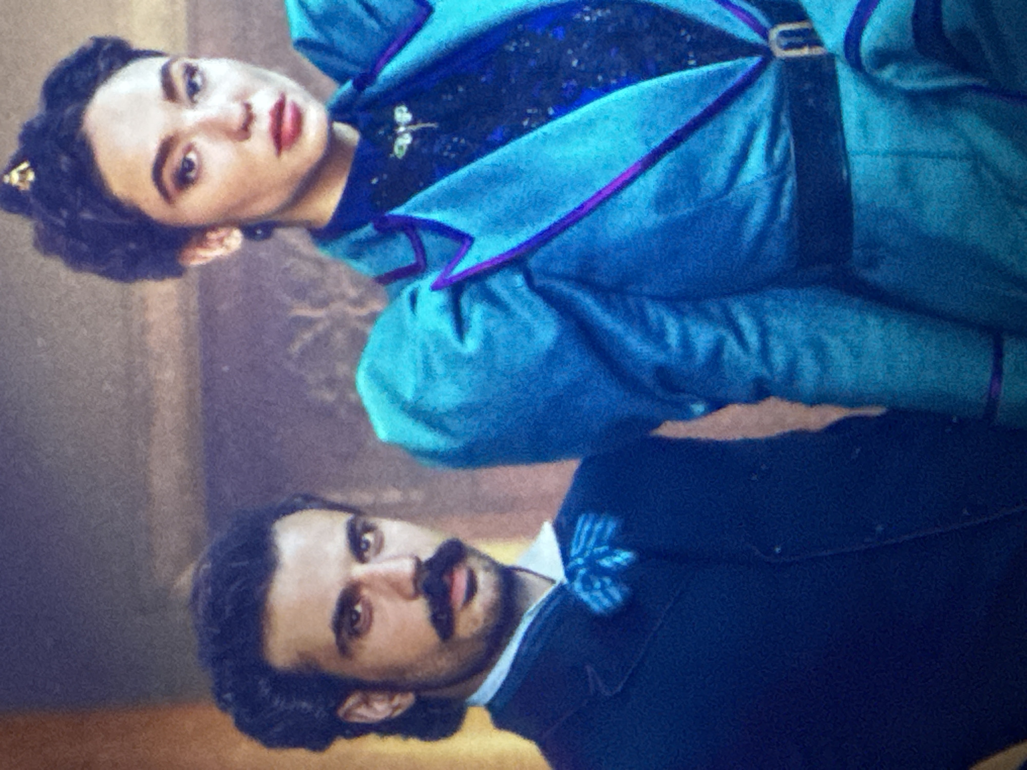 This media file displays two individuals captured in a portrait-style composition, oriented with a 90-degree clockwise rotation.
In the upper section of the rotated image, a woman with dark hair styled in an elaborate updo features a subtle golden accessory. She possesses fair skin, defined dark eyebrows, and red-painted lips, her gaze directed straight at the viewer with a neutral, perhaps serious, expression. She is attired in a vibrant teal-blue jacket accented with distinctive purple trim along its edges, worn over a dark, possibly sequined or intricately patterned, inner garment. A dark belt with a rectangular gold-toned buckle is visible at her waist.
Below her, a man with dark, curly hair, a prominent mustache, and a short beard also looks directly at the viewer with a steady, composed demeanor. He is dressed in a dark suit jacket, a white collared shirt, and a striped blue bow tie or cravat.
The background is a muted and textured surface, primarily in shades of brown and beige, giving the impression of an indoor setting, possibly a studio or a historically themed environment. Both subjects appear to be formally posed, suggesting a portrait sitting. The attire of both individuals suggests a period or stylized aesthetic. There is no visible text in the image. The context provided is Puebla, Mexico.