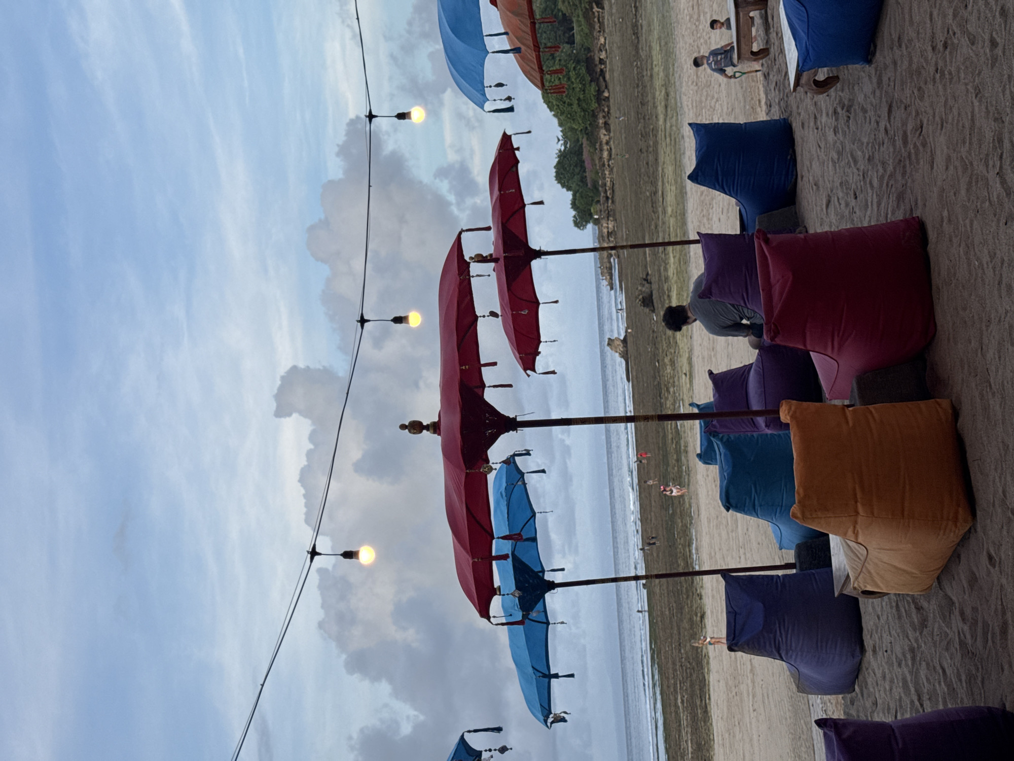 The image depicts a beachfront scene in Kuta Selatan, Indonesia, featuring a row of colorful beanbag chairs arranged on the sand. Overhead, a string of lights is strung between decorative umbrellas in shades of red, blue, and brown. In the background, a sandy beach stretches towards the ocean, with a line of vegetation visible in the distance. Several people can be seen on the beach and sitting in the beanbag chairs. The sky is cloudy.