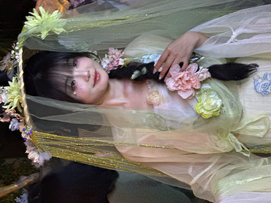 This image captures a close-up portrait of a young East Asian female, located in Yuzhong District, China, under artificial lighting in what appears to be an indoor or sheltered environment. The subject is depicted from the waist up, facing forward with a slight smile. She is dressed in a multi-layered, traditional Chinese-style garment, predominantly in light green and cream hues, extensively adorned with artificial flowers, including large pink peonies and smaller white and green blossoms. A pale blue embroidered floral design is present on the lower section of her dress. Her long, dark hair includes bangs, and a thick braid positioned over her right shoulder is decorated with additional flowers. She wears a wide-brimmed woven hat, accessorized with various flowers, a translucent light green veil, and shimmering golden tinsel strands. Her facial makeup includes glitter on the eyelids and cheeks, with small pink heart-shaped gems situated beneath her eyes. Her left hand, displaying long, manicured nails with an iridescent polish, is gently placed on her decorated braid. The background is dark and out of focus.