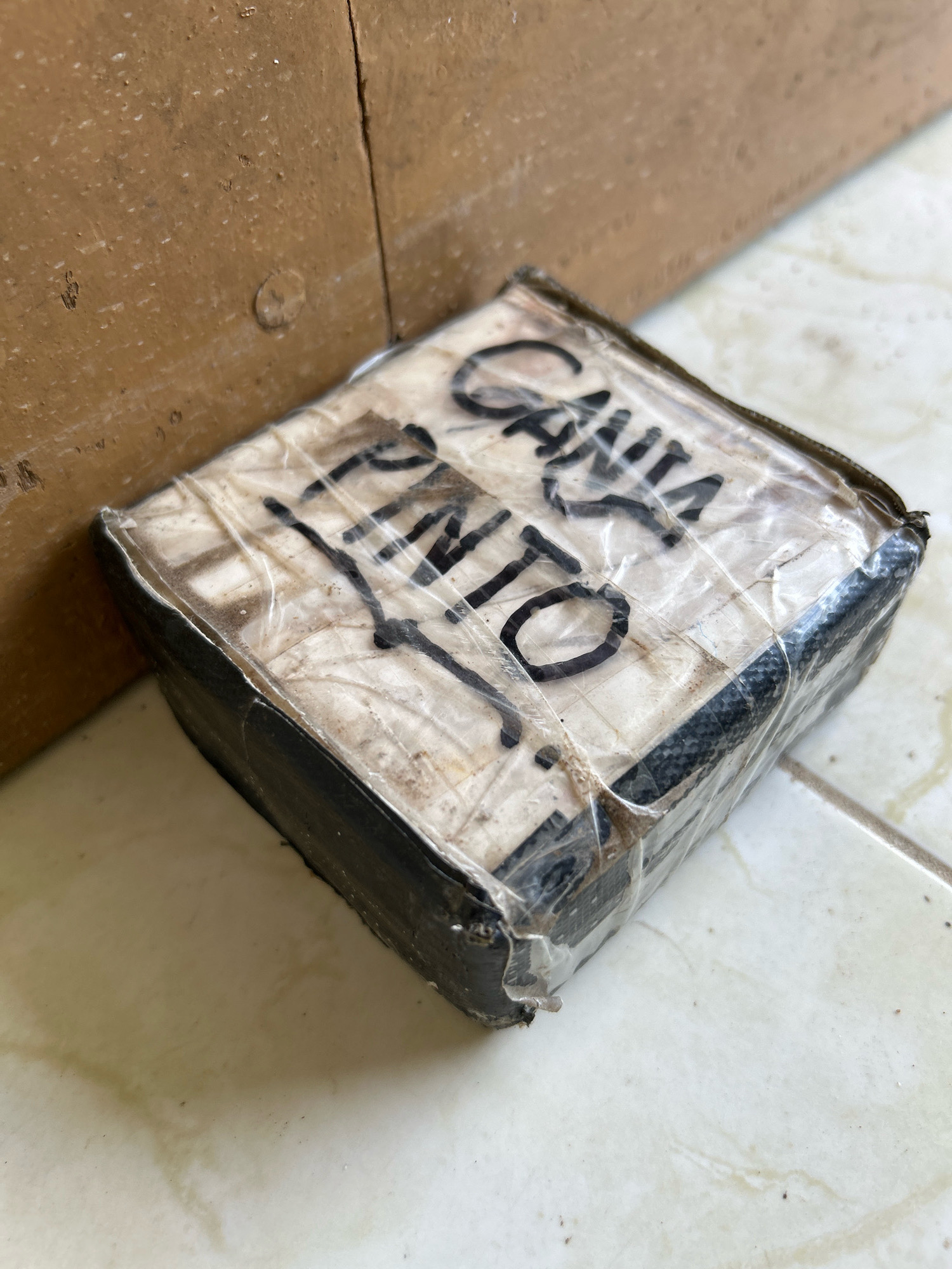 A small, rectangular package wrapped in plastic wrap and black tape is resting on a tiled floor next to a cardboard box. The package has writing in black marker that appears to say "CANVA PINTO." The scene is outdoors, with the tiled floor and a hint of a wall in the background. The lighting suggests it is daytime. The overall condition of the package suggests it may have been handled or shipped. There are no people visible in the image. The location is described as the city of Sabang, Indonesia.