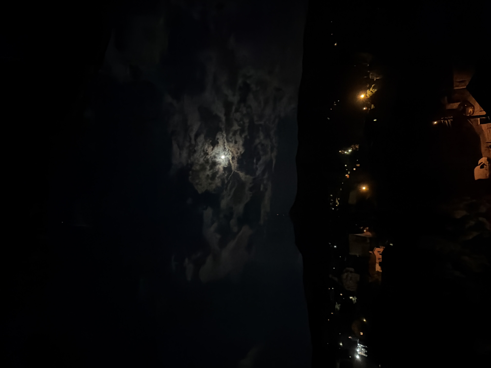 The image shows a night scene with a bright moon surrounded by clouds in the left half of the frame. The right half is darker, showing a residential area with street lights and some buildings visible. It is too dark to see any people. The location is in Sofia, Bulgaria.