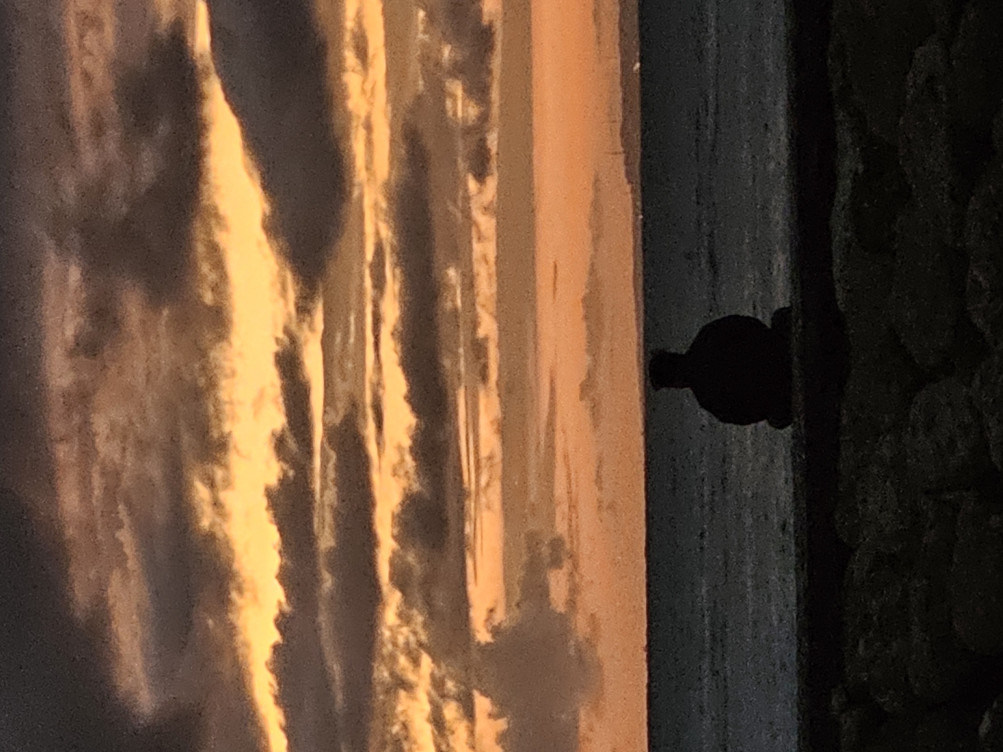 A silhouette of a person sits facing the ocean, with their back to the viewer. The person is seated on a dark, rocky surface, with their knees bent and their arms resting on their knees. The ocean stretches out before them, with a hint of a distant shoreline visible on the horizon. The sky above is filled with dramatic clouds, illuminated by the warm hues of a sunset or sunrise. The colors range from deep oranges and yellows to darker grays and purples, creating a striking contrast between the light and shadow. The overall mood of the image is one of quiet contemplation and solitude. The scene is set in Denpasar, Indonesia, a city known for its beautiful beaches and vibrant sunsets.