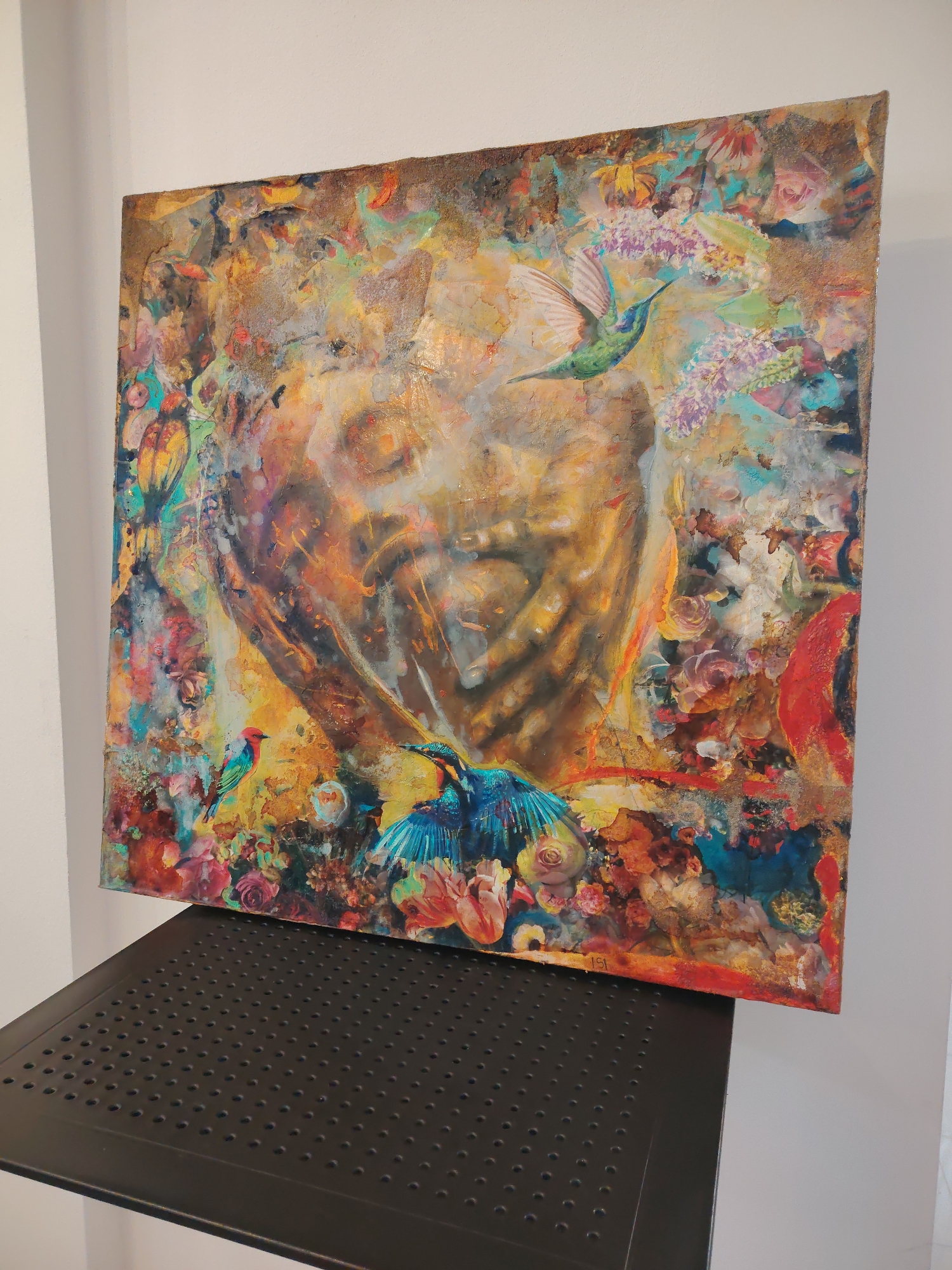 A square painting rests on a dark, perforated surface. The painting is vibrant, with a central, blurred figure of a face with hands near the mouth and chin. The face is rendered in hues of brown, orange, and yellow. Surrounding the face are flowers, foliage, and birds. A hummingbird is depicted near the upper right quadrant of the image. The edges of the painting are textured and appear to be a mix of brown and gold. The painting is positioned near a light-colored wall. The location is not discernible from the visual information provided. There is no indication of the city of Varna, Bulgaria.