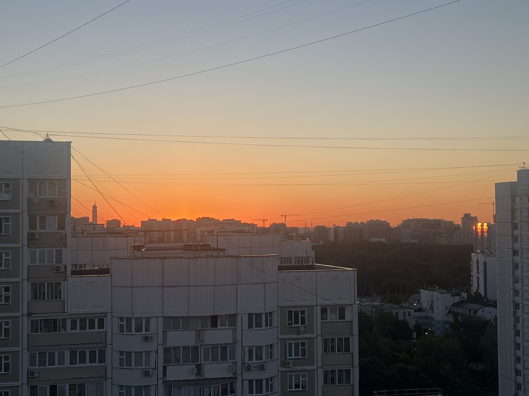 The image captures a sunset over a densely populated urban area in Moscow, Russia. The sky transitions from a pale blue at the top to a vibrant orange and yellow hue closer to the horizon, indicative of the setting sun. Several high-rise apartment buildings, characteristic of Russian residential architecture, dominate the foreground and midground. These buildings are constructed with concrete panels, featuring numerous windows and balconies. Power lines crisscross the sky above the cityscape. In the distance, more buildings and some construction cranes are visible, silhouetted against the bright sky. A dense patch of trees can be seen between the midground and background buildings. The setting sun is partially obscured by the buildings, casting a warm, golden glow and reflecting off some of the windows. There are no people visible in the frame. The overall scene depicts a typical evening in a Moscow residential district.