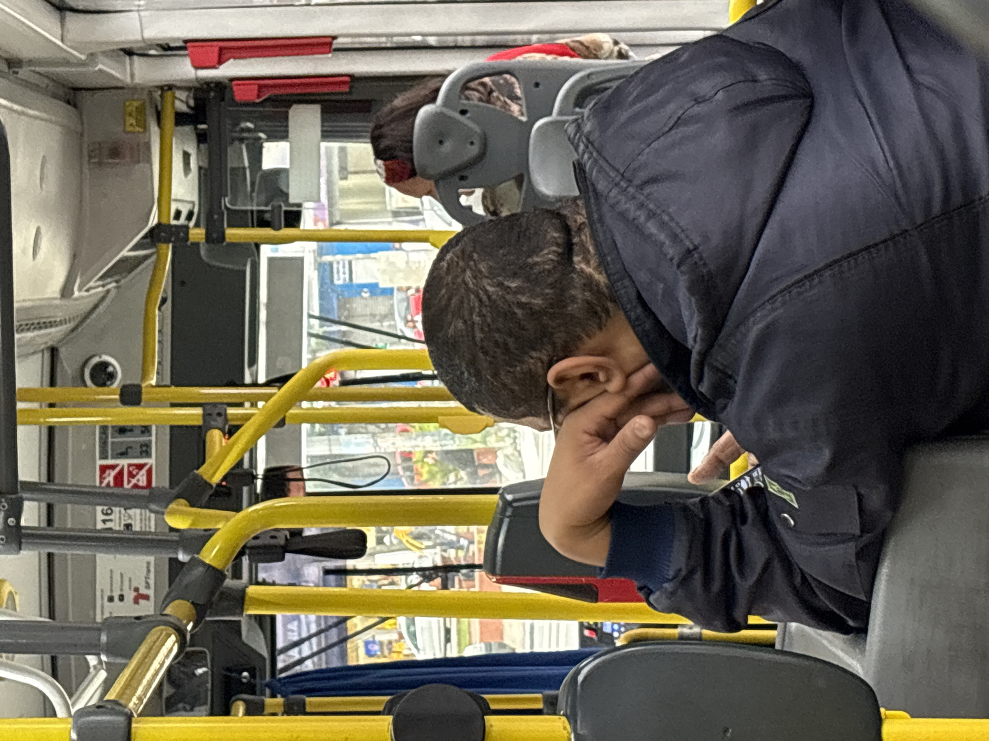 The image shows the interior of a public bus in São Paulo, Brazil. In the foreground, a person wearing a dark blue jacket is seated, with their head resting on their hand and arm. The person is facing away from the camera, their gaze directed forward and slightly downward. The bus interior features yellow poles, gray seats, and some red handrails. Through the windows, glimpses of the city can be seen, including buildings and other vehicles, indicating an urban environment. Another person with long dark hair, wearing a red garment, is seated in front of the person in the foreground. The overall scene captures a moment of quiet travel on a bus.
