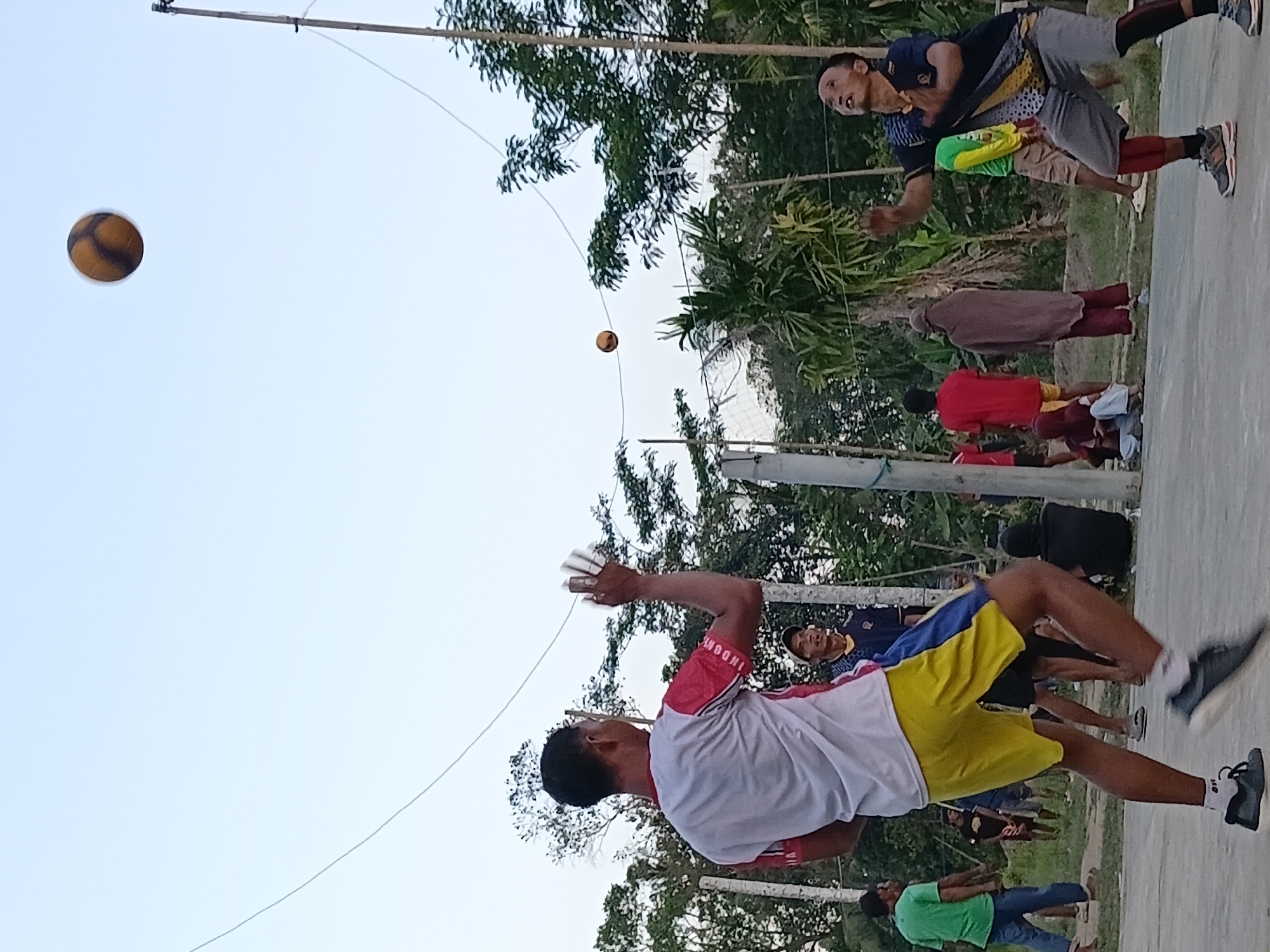 In Margo Rukun, Indonesia, a group of people are engaged in a game of volleyball outdoors on a court. The sky is clear and light blue, indicating it is likely daytime. In the foreground, two men are actively playing. One man, wearing a white and red shirt and yellow shorts, is in the process of serving or hitting the ball, with his left arm raised and holding what appears to be a shuttlecock or a small ball. His right arm is extended, and his body is in motion. He is wearing black and white athletic shoes with white socks. The other active player, wearing a dark blue shirt with yellow and black accents and grey shorts, is positioned on the right side of the court and is looking up, preparing to receive or hit the ball. He is wearing athletic shoes and knee-high socks.

A volleyball is visible in the air, high above the players. Another smaller ball, possibly a shuttlecock, is also in the air. In the background, several other individuals are observing the game or participating in other activities. A man in a green shirt and blue jeans stands to the left, facing the court. Further back, a woman in a brown headscarf and maroon clothing is standing. Other figures, some seated and some standing, are scattered around the periphery of the court. The court itself appears to be made of concrete. A volleyball net is set up, and poles support it, with wires strung overhead. Lush green trees and foliage surround the court, creating an outdoor, natural setting.