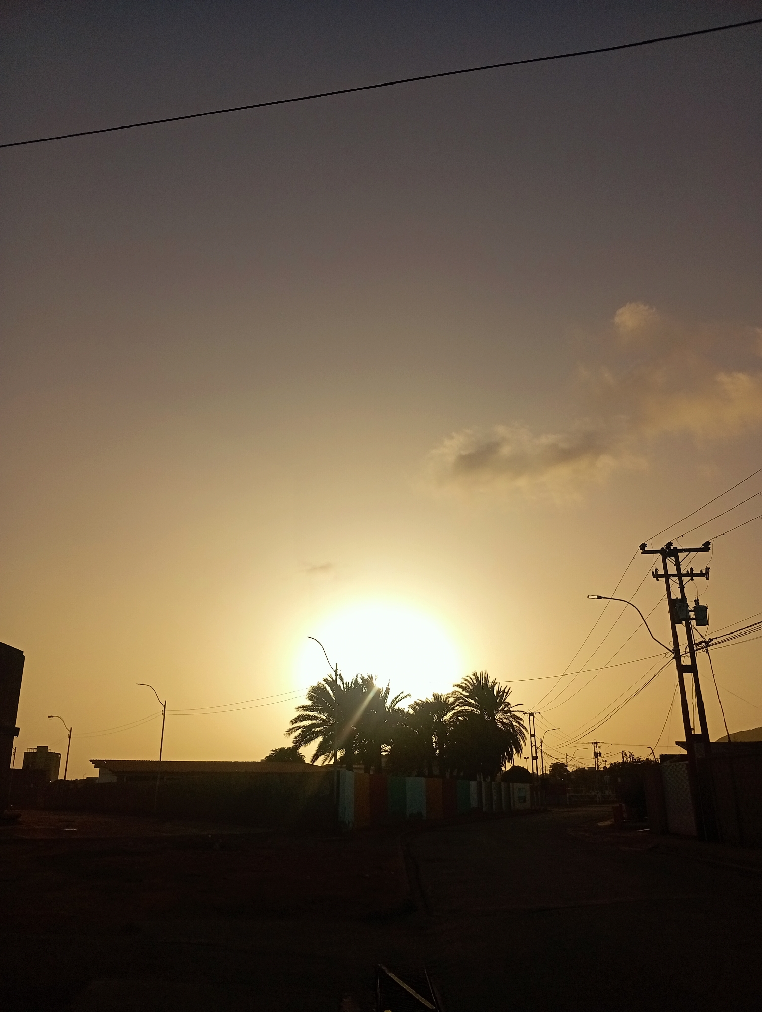 The image depicts a serene sunrise or sunset over a street in Juan Griego, Venezuela. The sky presents a dramatic gradient, shifting from a dark grey-purple at the top to an intense golden-orange hue centered around the sun. The sun itself is a brilliant, glowing orb, partially obscured by the silhouetted fronds of several palm trees located in the middle ground. A few scattered clouds in the upper sky are faintly illuminated by the warm light.
In the foreground and middle ground, the landscape is rendered in dark silhouettes against the bright sky. A prominent utility pole with multiple power lines stands on the right side of the frame, with wires stretching across the upper portion of the image. Two streetlights are also visible as dark silhouettes. Along the seemingly empty street, the outlines of low-rise buildings, walls, and fences can be seen. Behind the palm trees, a section of a wall or fence features colorful vertical stripes. Faint outlines of distant hills or mountains are visible in the far background on the right. The overall impression is one of quiet transition, with the natural beauty of the sky contrasting with the man-made infrastructure. There is no visible text or explicit activity taking place.