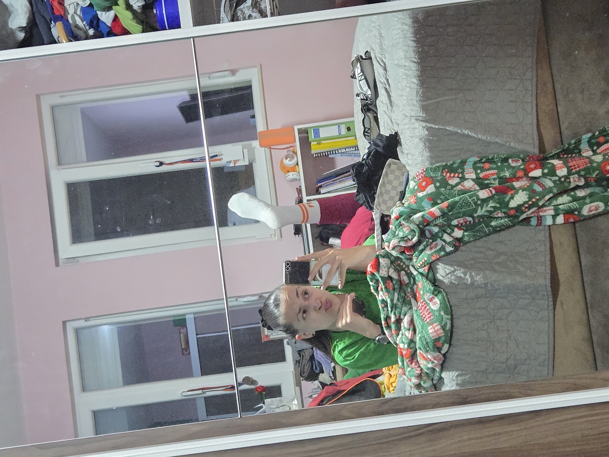 A person is taking a selfie in a mirror. The reflection shows a room with a pink wall, two windows, and a bed with a patterned blanket. The person is wearing a green shirt, orange and white striped socks and red pants. There is a white shelf with books and a stuffed animal. The location appears to be indoors. There is not enough information to determine the location in the city of Pleven, Bulgaria.