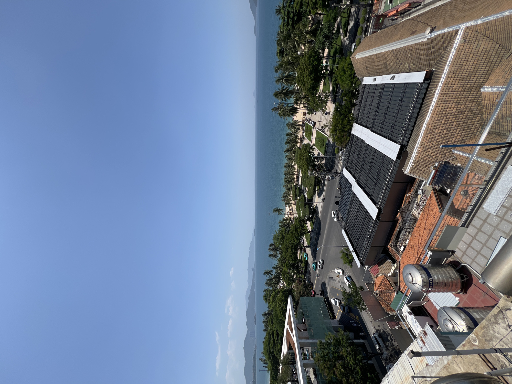 The image captures a panoramic view of a coastal city, likely Nha Trang, Vietnam, on a clear, sunny day. The foreground features several rooftops with various materials, including corrugated metal and terracotta tiles, as well as large cylindrical water tanks, some appearing to be stainless steel. A metal railing is visible in the lower right corner.
Beyond the rooftops, a street is visible with several cars driving. Adjacent to the street is a landscaped area with trees and a pedestrian walkway leading to a beach. The beach is lined with palm trees and appears to have people present, though they are small in scale.
The sea, a deep blue, stretches to the horizon, meeting a clear, light blue sky. Distant mountains form a hazy backdrop along the coastline. The overall scene depicts a typical urban coastal environment with a blend of residential, commercial, and recreational elements. The lighting suggests it is midday. No specific individuals or actions are discernible in detail due to the distance.
