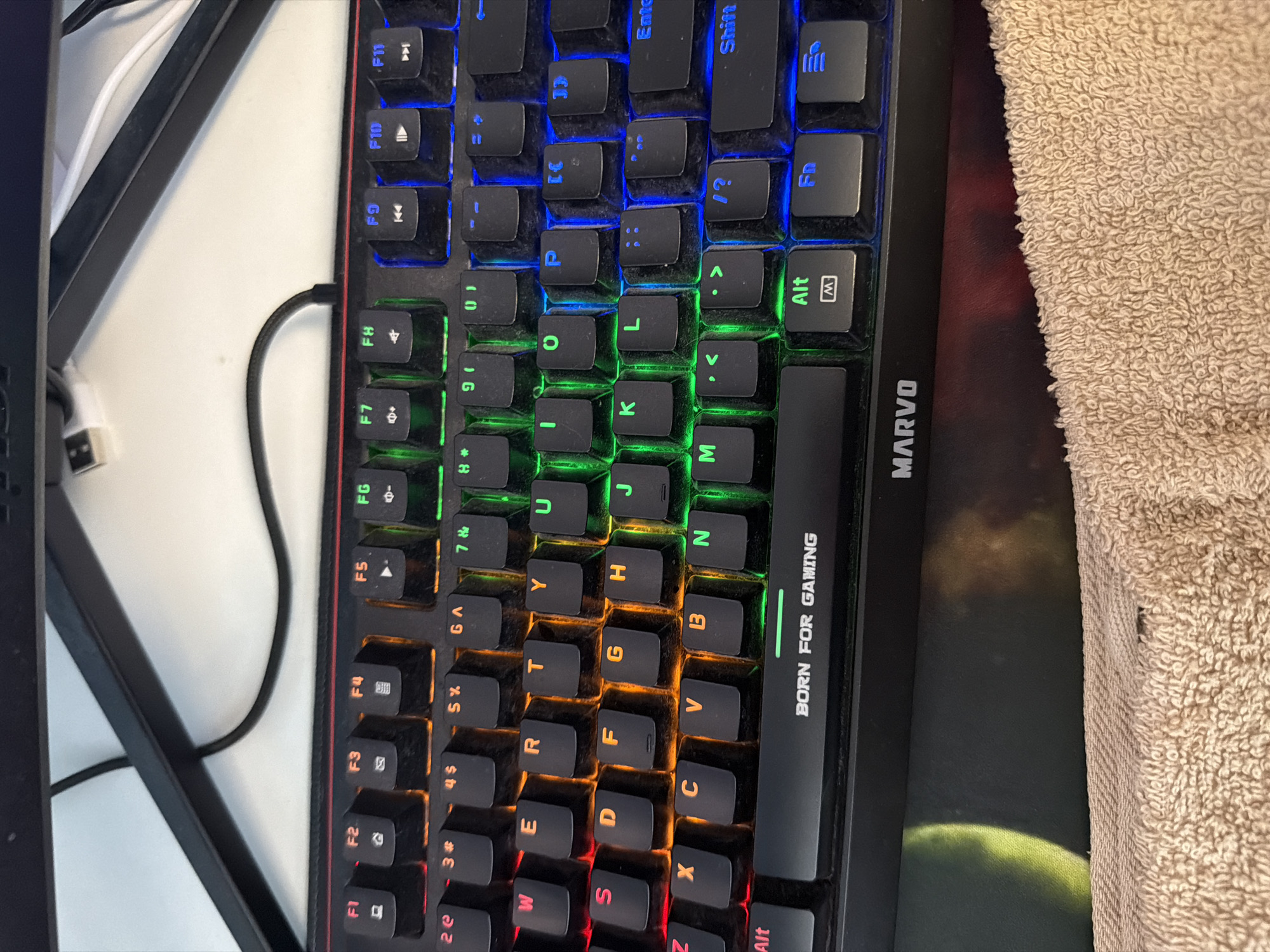 An overhead shot captures a gaming keyboard, mousepad, and a light-colored towel on what appears to be a desk or table. The mechanical keyboard is the central subject, featuring multi-zone RGB backlighting that transitions from blue at the top (function keys, number row) to green (QWERTY home row), orange (middle alphanumeric rows), and red (bottom rows). The keys are visibly dusty.
The keyboard is a QWERTY layout with standard alphanumeric keys, function keys (F1-F12), media control icons, and modifier keys like Alt, Shift, Ctrl, and an Fn key. The right side of the keyboard's frame displays the brand name "MARVO" vertically, with the slogan "BORN FOR GAMING" directly below it. A black braided cable, likely connecting the keyboard, is visible extending towards the upper left.
Partially visible beneath the keyboard and a bunched-up, light beige terry cloth towel on the right is a mousepad. The mousepad also bears the "MARVO" brand name and features a dark background with a hazy, green or yellow-green globe-like graphic on the lower section. In the upper left corner of the image, sections of white cables and a dark monitor stand or arm are visible against a plain white surface, suggesting a typical computer workstation setup.
