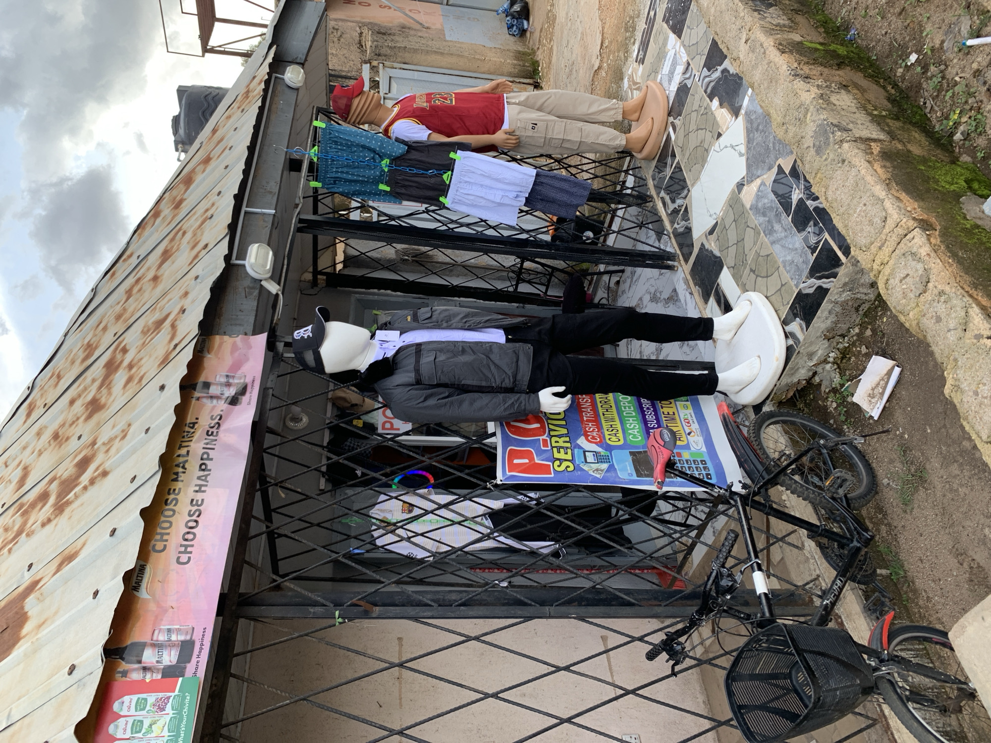 This media file shows an outdoor market stall in Jos, Nigeria. Two mannequins are displayed in front of the stall. The mannequin on the left is wearing a gray jacket, white shirt, and black pants, with a white cap. The mannequin on the right is wearing a red vest with the number 23, a white t-shirt, beige cargo pants, and tan shoes. Several pairs of shorts are hanging on a rack between the mannequins.
A bicycle with a black basket is parked next to the stall. In the background, there is a corrugated metal roof and a building with tiled walls. A large banner advertising "MALTINA" and "CHOOSE MALTINA. CHOOSE HAPPINESS." is attached to the side of the stall. Another sign displays services like "CASH TRANSFER," "CASH WITHDRAW," and "CASH DEPO."
The sky is overcast, suggesting it may be a cloudy day. The overall scene depicts a typical street vendor setup in a Nigerian urban environment, with clothing and advertising displayed to attract customers. The details suggest a commercial activity taking place at what appears to be daytime.
