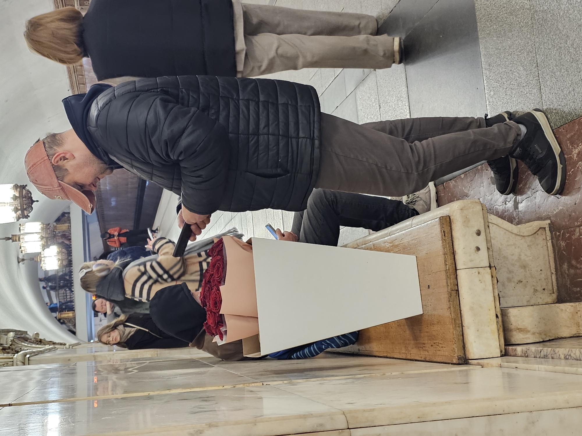 The image depicts a scene inside a Moscow Metro station in Russia, showing several people and objects.
A man wearing a black quilted jacket, brown trousers, and black sneakers is standing. He's holding a black object in his hand and looking towards a large bouquet of red roses in a tall white box. A woman with blonde hair is standing to his left, wearing a black vest and beige trousers. Several other people are seated on a wooden and marble bench, some of whom appear to be looking at their phones.
The environment is a typical Moscow Metro station with ornate decorations, including chandeliers and patterned walls. The floor is made of marble. Overall, the scene captures a moment of everyday life within the context of the city's subway system.