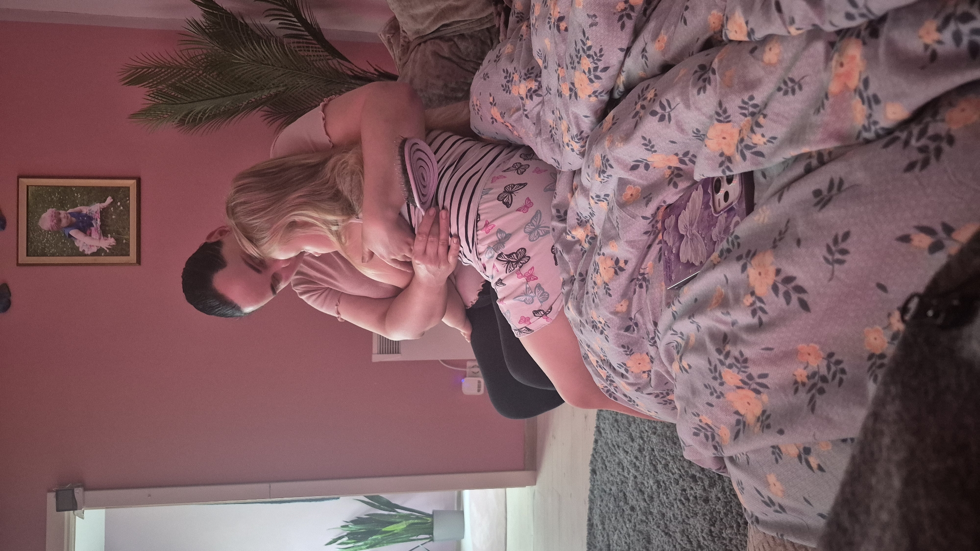 The image depicts an indoor scene, likely a bedroom, with a person holding a child. The room has pink walls, and a framed picture of a child is visible on the wall. There is a plant in the corner and a radiator. The person is wearing a pink shirt and dark pants, and the child is in a striped top and shorts with a butterfly pattern. They are sitting on a bed with floral bedding. A hairbrush is being held by the person, and a phone is visible on the bed. The overall impression is one of a tender moment between the two individuals. The scene is not specifically identifiable as being in Švenčionys, Lithuania, as there are no unique landmarks or contextual clues to suggest this location.