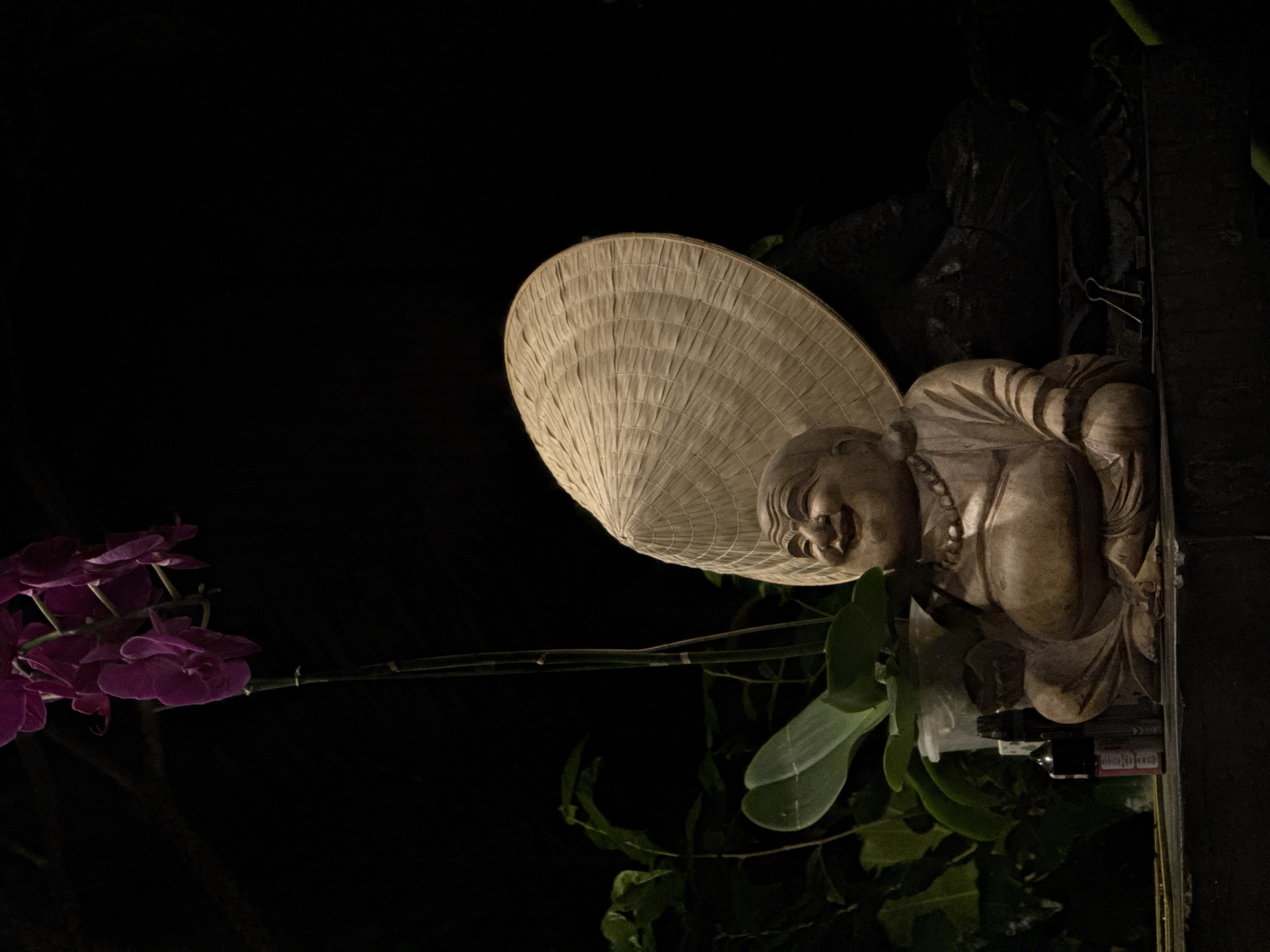 The image shows a carved wooden statue of a laughing Buddha wearing a conical straw hat. The statue is positioned vertically, facing towards the left of the frame, with its head tilted down. To the left of the statue, a branch of vibrant purple orchids is in bloom. The background is dark, suggesting it is nighttime or a dimly lit area. Green foliage is visible behind the statue and the orchids, partially obscuring the background. The statue appears to be outdoors, possibly in a garden or courtyard. A small black object, possibly a light or electronic device, is attached to the right side of the statue. There are no visible people or any clear indication of weather. The overall mood is peaceful and serene, with the bright flowers providing a pop of color against the dark backdrop and the statue's warm wooden tone. No text is visible in the image.