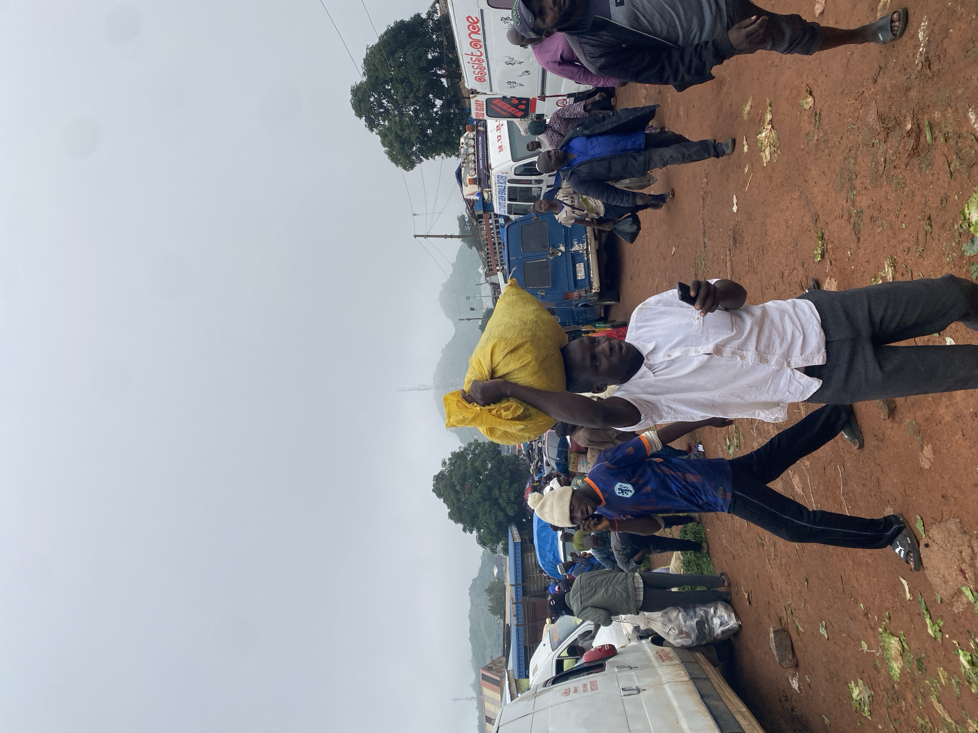 A street scene in the city of Katon Rikkos, Nigeria, shows a busy marketplace on a cloudy day. In the foreground, a man with a white shirt and grey pants carries a large yellow bag on his head. He is holding a black smartphone in his left hand and looking down at it. To his left, a man in a blue and orange soccer jersey and a white beanie is talking on a black smartphone. Further back, a white van is parked on the left side of the frame, with a large bag of produce next to it. Several other vans and people are visible in the midground and background, suggesting a bustling commercial area. The ground is covered in reddish-brown dirt, with some scattered green leaves. In the distance, green hills are visible through the haze. The overall impression is one of a vibrant, active community engaged in daily commerce.
