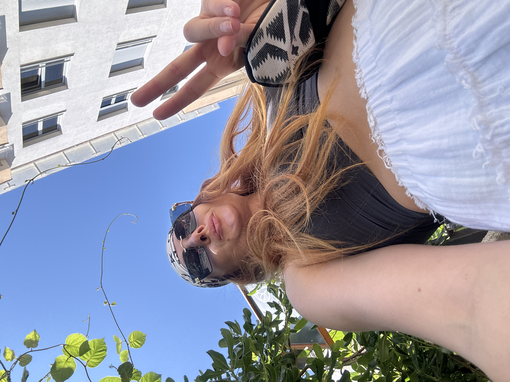 A woman with long, red hair, wearing a black top, a white ruffled skirt, sunglasses, and a patterned headscarf, poses for a selfie outdoors. She is looking up at the camera and making a peace sign with her right hand. In the background, a white building with windows and a clear blue sky are visible. Green foliage is also present on the left side of the image. The location is identified as Budva, Montenegro.