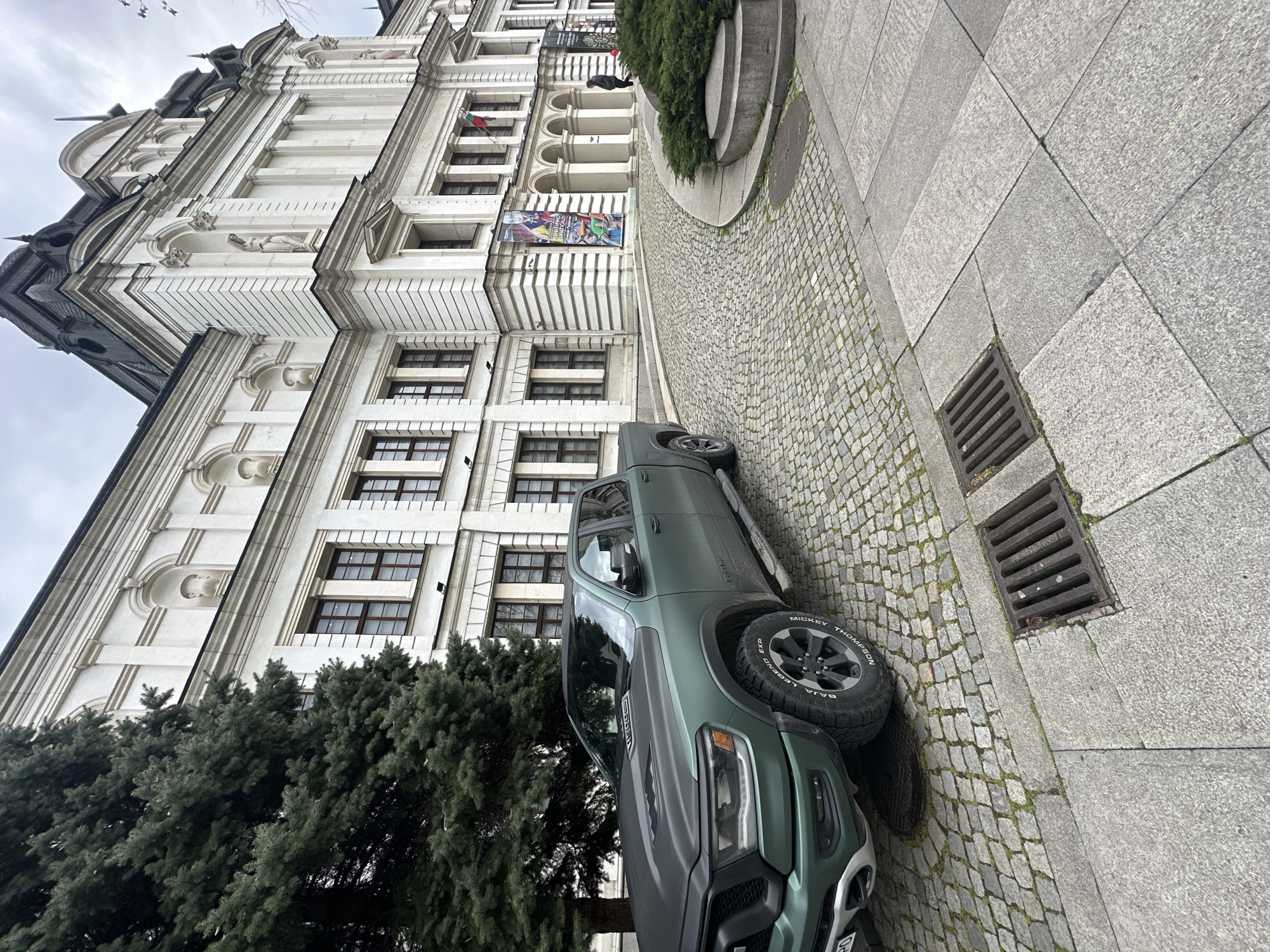 An outdoor, slightly tilted shot captures a grand, multi-story building in Sofia, Bulgaria, alongside a green pickup truck and a cobbled street on an overcast day.
The prominent structure, likely a historical or official building, features a light-colored, ornate facade with classical architectural elements. It has multiple windows with dark frames, several balconies, and decorative stone carvings including sculptural figures within niches. A dark, steeply pitched roof with spires or decorative elements is visible at the top. Near one of the building's arched entrances, a person in dark clothing stands. Two banners hang from the facade: one is colorful and abstract, while another displays the Bulgarian flag.
In the foreground, a dark green pickup truck, specifically a RAM 1500 (indicated by text on the hood), is parked on a cobblestone street. Its tires, manufactured by "Mickey Thompson," feature the "Baja Legend EXP" model name. The street transitions from small cobblestones near the truck to larger, grey square paving slabs. Several metal drain grates are set into the ground. Lush, dark green coniferous trees are visible on the left side of the frame, partially obscuring the lower part of the building and the front of the truck. The sky above is bright and cloudy.