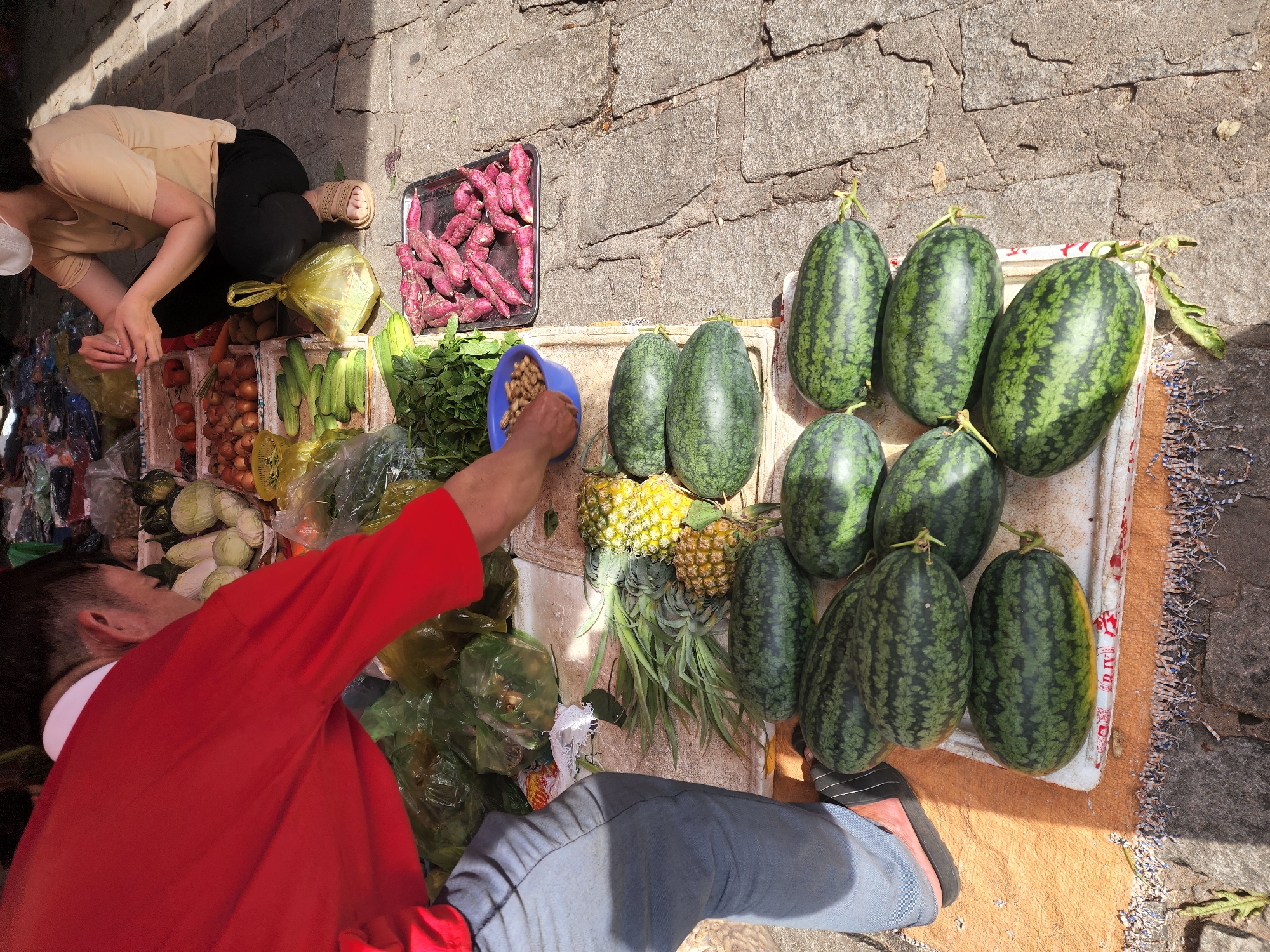 An overhead shot captures a street market scene in Phu Thuy Ward, Vietnam. A man in a red shirt and blue jeans is reaching into a blue bowl, his hand submerged, as he interacts with a stall displaying various produce. To his right, a woman wearing a light-colored t-shirt and dark pants is crouched down, her face obscured by a white mask, and she is looking at her hands. The stall features an assortment of fresh vegetables and fruits, including watermelons, pineapples, sweet potatoes, and what appear to be cucumbers. The ground is paved with rough stones.