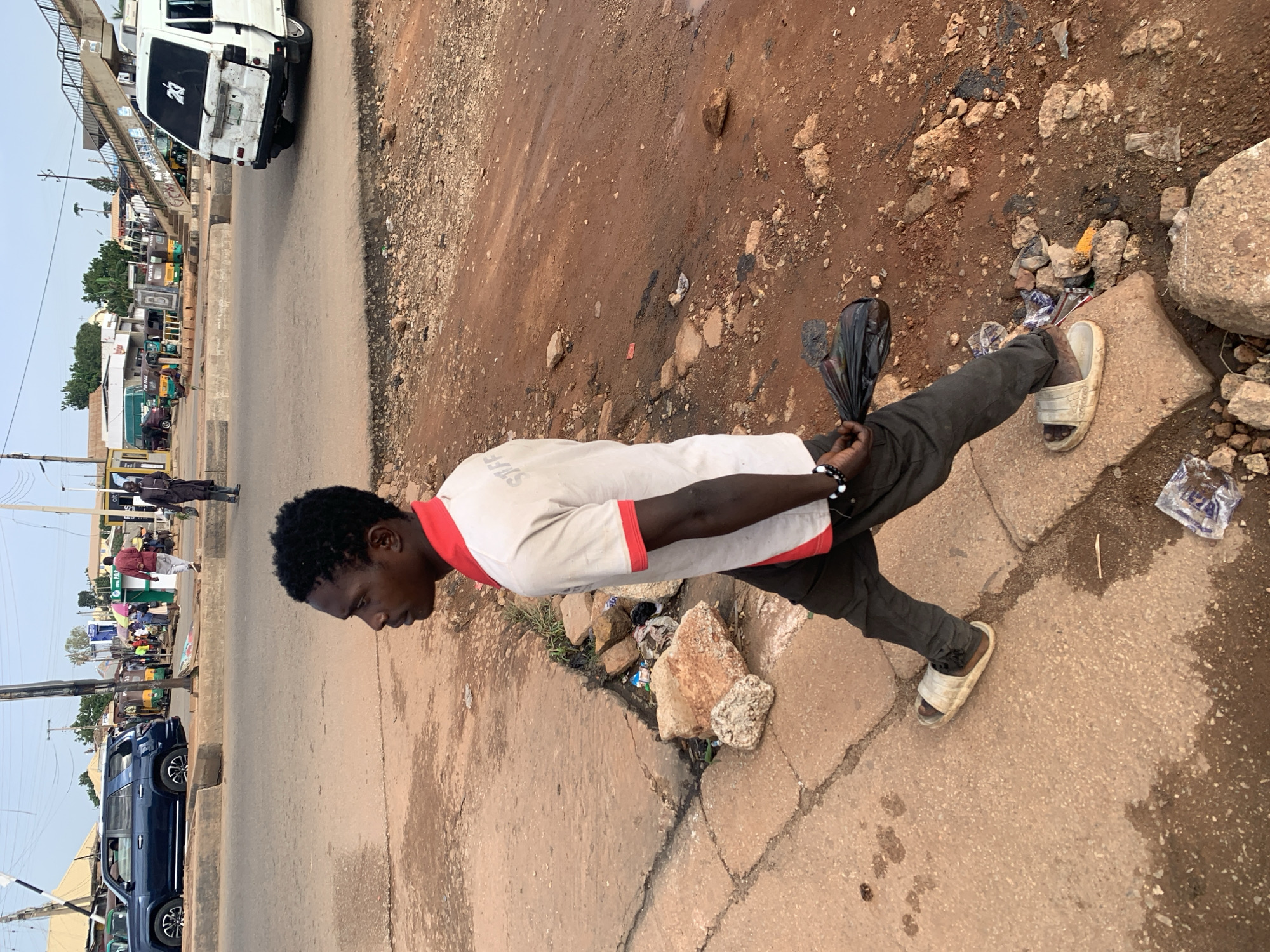 A young Black male, wearing a white shirt with red trim and dark pants, walks along a dirt and concrete path in Gura Top, Nigeria. He is holding a black plastic bag in his right hand and appears to be wearing wristbands on his left hand. The path is uneven and littered with rocks and debris. In the background, a road is visible with several vehicles, including a dark blue SUV, a white van, and various colorful tricycles, commonly known as Keke Napeps. Pedestrians are also present in the background, along with buildings and a pedestrian overpass. The overall scene suggests a busy urban environment with signs of commercial activity and everyday life.