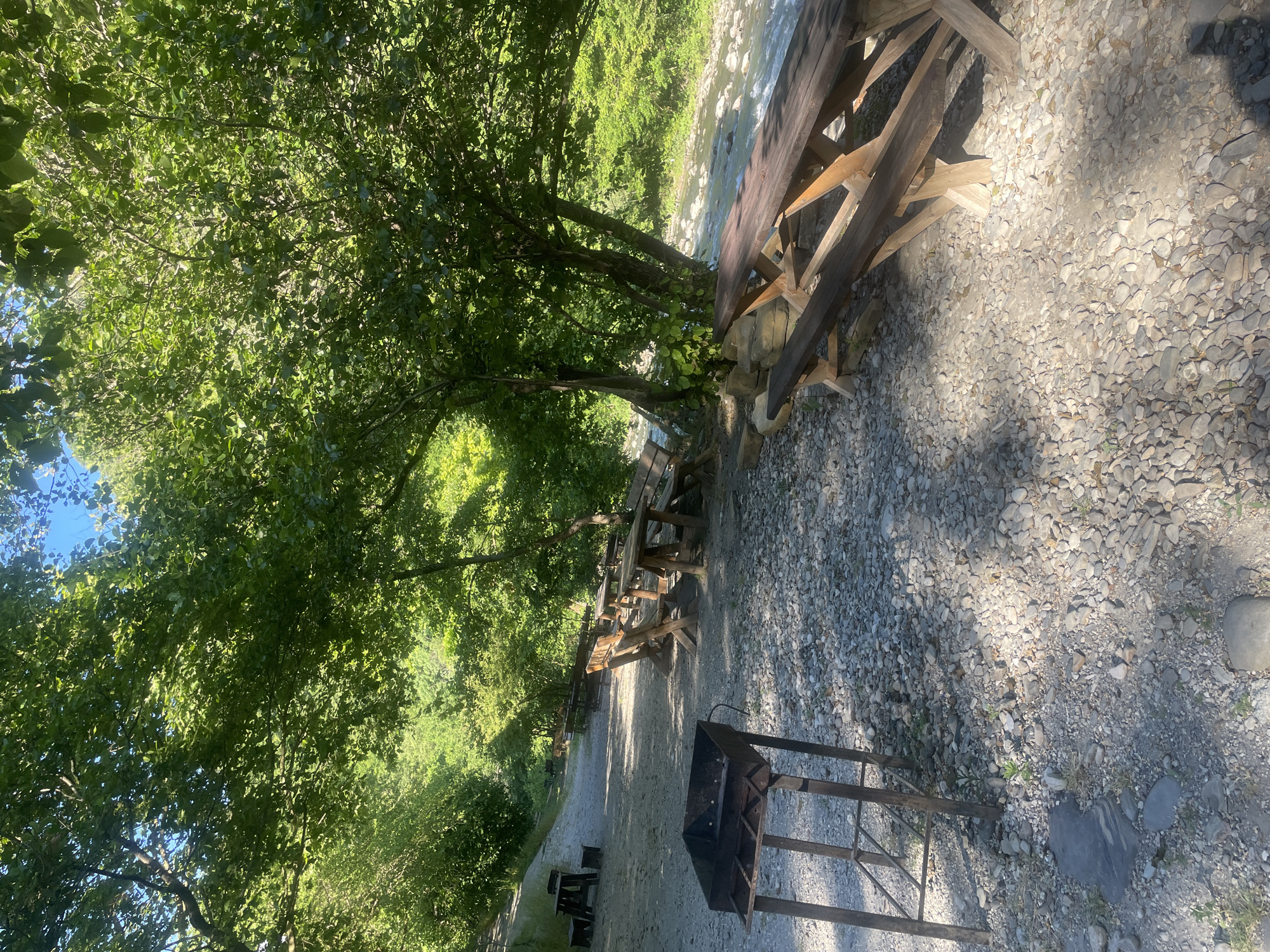 The image depicts a serene outdoor picnic area situated by a flowing river, likely within the city of Sochi, Russia. The scene is dominated by lush green trees providing ample shade, with sunlight filtering through the leaves, casting dappled shadows on the pebbled ground.
In the foreground, a rustic metal barbecue grill stands on four legs, suggesting readiness for use. Several wooden picnic tables with attached benches are arranged along a gravel path, some of which are positioned closer to the riverbank, offering scenic views. The tables and benches appear sturdy and well-maintained, indicating a functional recreational spot.
The river itself is visible in the background, its clear water cascading over rocks, creating a gentle, natural ambiance. The surrounding environment is characterized by verdant foliage, characteristic of a forested or park-like setting. The overall impression is one of a tranquil and inviting space for outdoor gatherings and relaxation, consistent with the natural beauty often found in the outskirts of Sochi. There are no people present in the image.