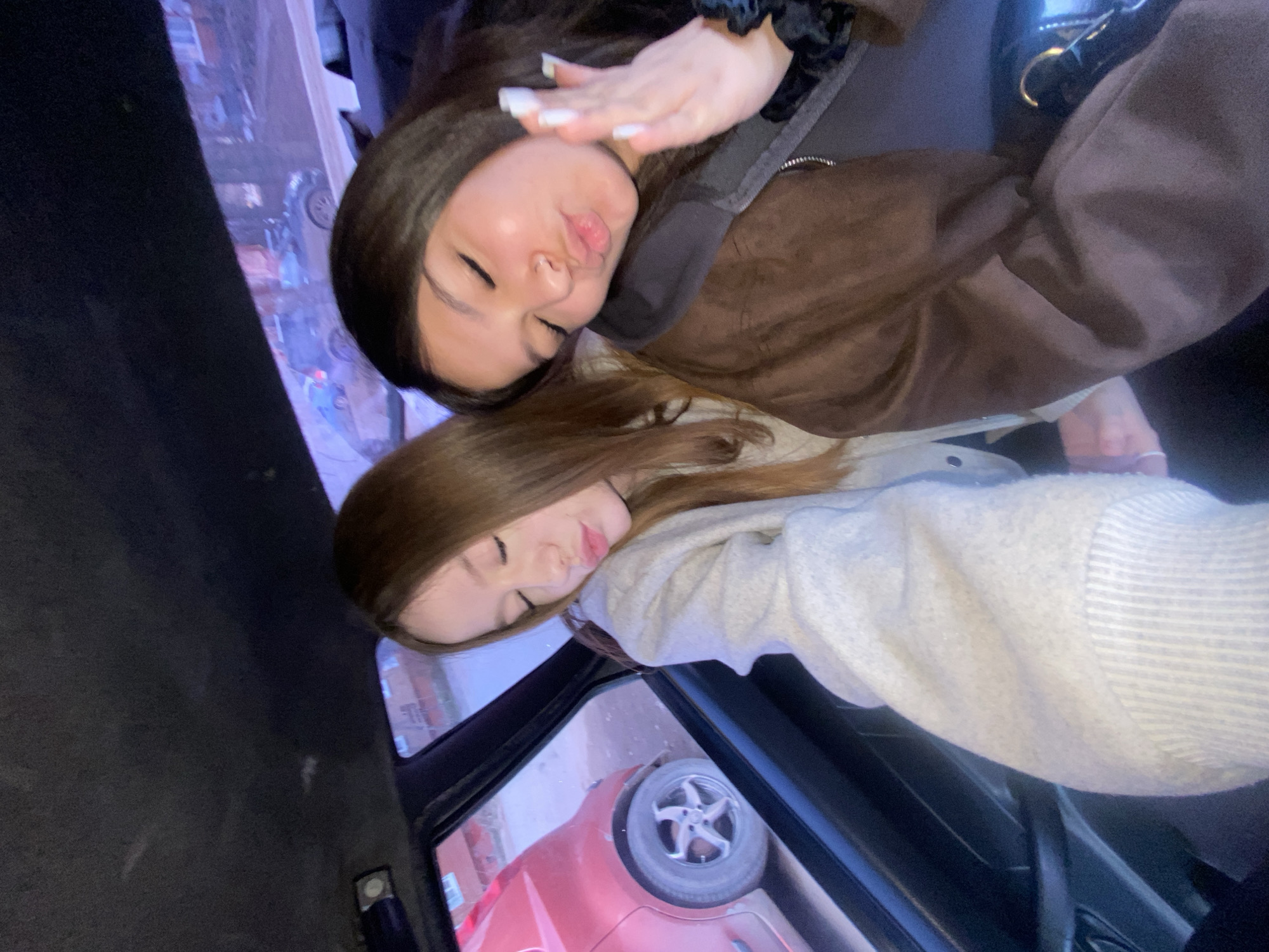 Two young women are photographed inside a car. One is lying on top of the other. The woman on top has dark brown hair and is wearing a brown jacket. She is making a kissy face and holding her hand up. The woman underneath has light brown hair and is wearing a light grey sweatshirt. Through the car window, a red car and building can be seen. The exact location within Votkinsk, Russia is not discernible from the image.