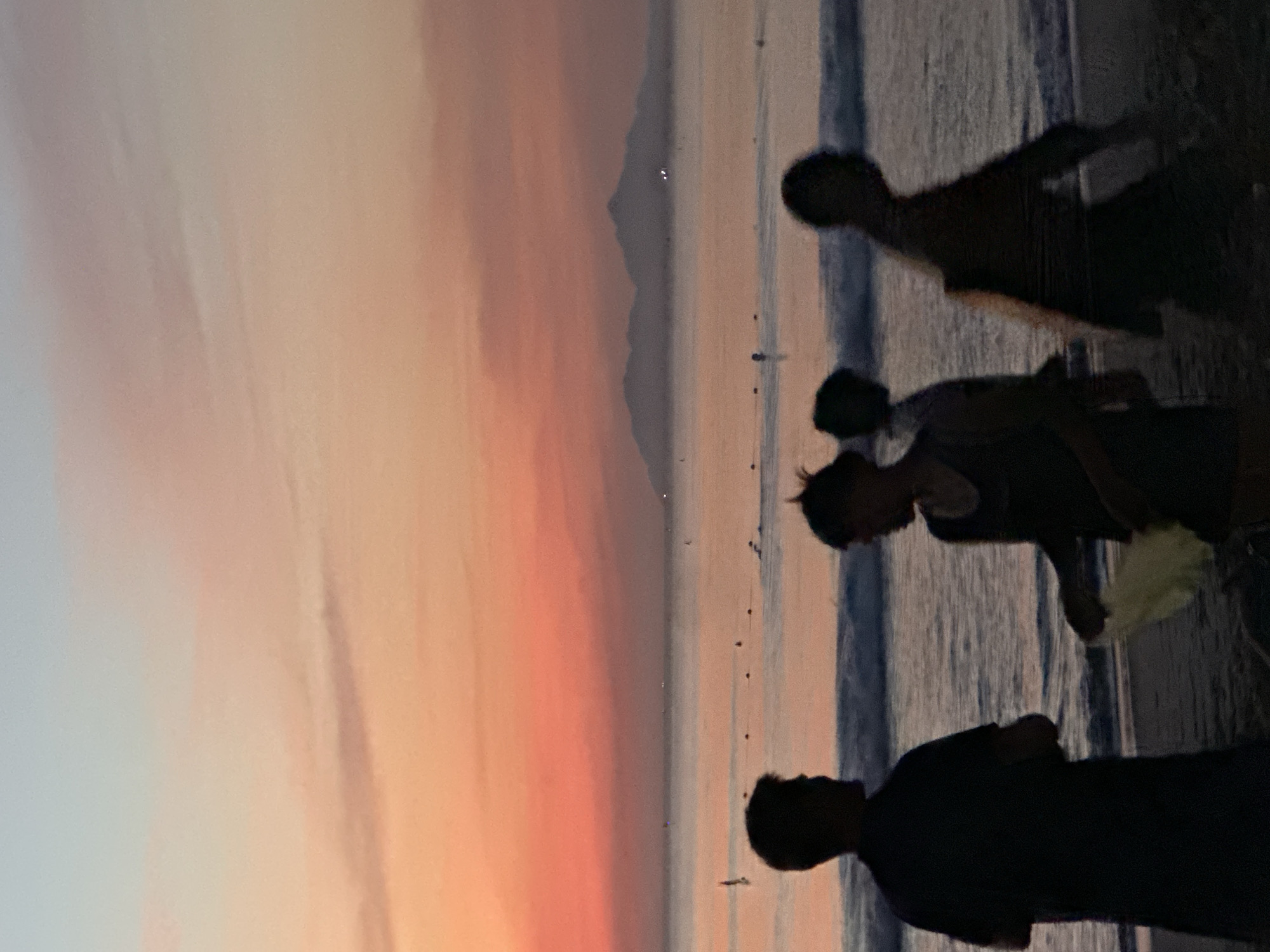 A silhouette of three people is visible on a beach at sunset. The sky is filled with hues of pink and orange, with a pale blue at the top. In the distance, a chain of mountains is visible across the calm sea, with a few small lights dotting the water, possibly from boats. Waves are gently rolling onto the shore. One person, on the left, stands facing the sea, their back to the camera. Another person, in the center, is facing slightly to the right, holding a light-colored object. The third person, on the right, is walking away from the camera, their form blurred by motion. The location is likely My Khe Beach in Da Nang, Vietnam, given the distinctive mountain range visible in the distance.