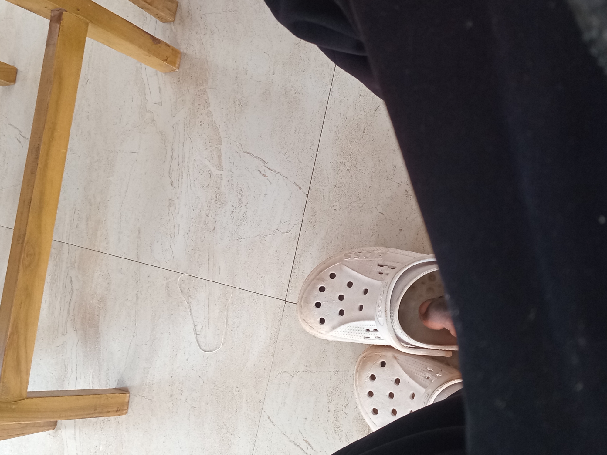 A close-up, overhead shot shows a pair of white Crocs on a person's feet, resting on a light-colored tiled floor. The person is wearing dark pants. The Crocs appear somewhat dirty and worn. In the background, the wooden legs of a chair are visible, suggesting the scene is indoors or on a covered patio. A thin white string lies on the floor between the chair legs and the Crocs. The environment suggests a casual setting, possibly in a home or a restaurant. The scene is devoid of any identifying markers of Bukuru, Nigeria.
