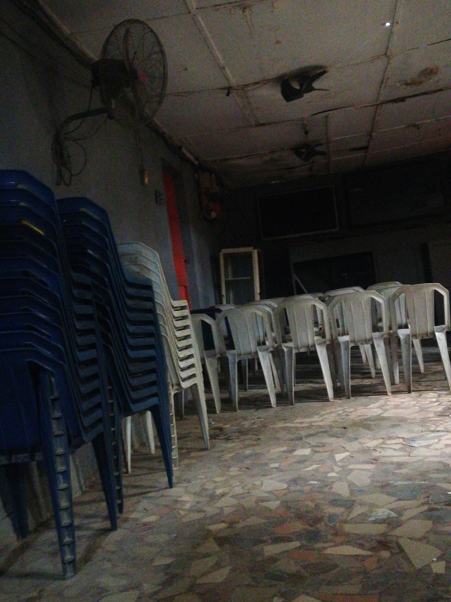 The image depicts an interior space in Lagos, Nigeria. The room is dimly lit with a visible ceiling fan, and stacked plastic chairs are arranged along the walls and in rows. The floor is tiled with fragmented patterns. A cabinet is visible towards the back, and a dark object hangs from the ceiling.