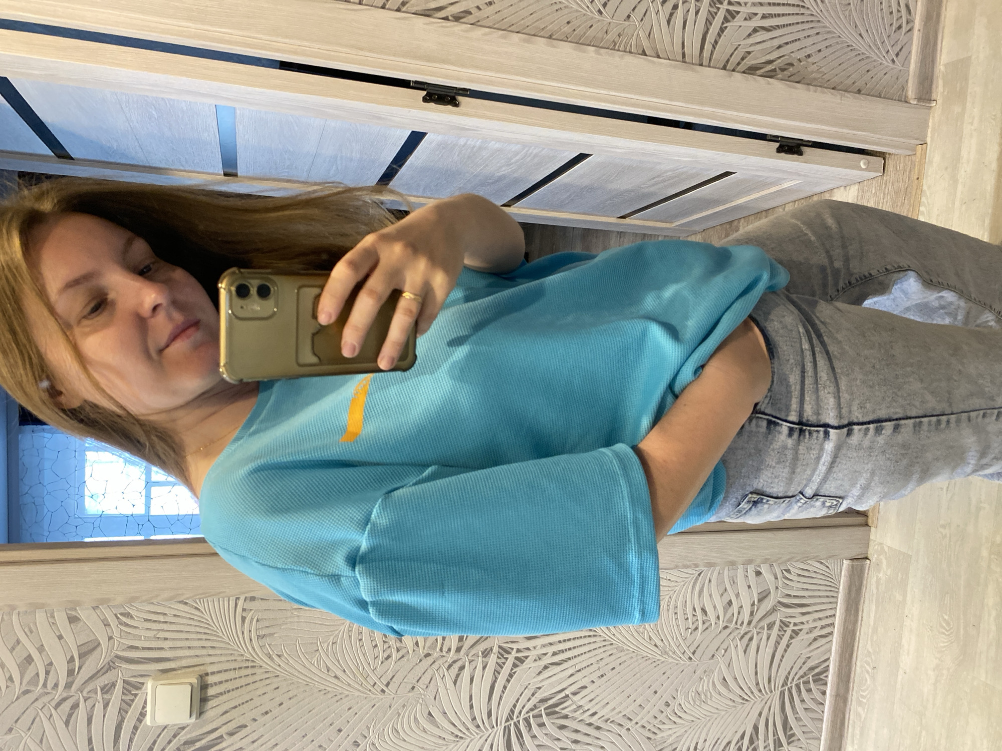 A woman is taking a selfie indoors. She is wearing a blue top with an orange stripe and light blue jeans. She holds a gold phone in her hand. The background includes a light-colored door, a wall with a palm leaf pattern, and a view of a window. The scene appears to be in an interior setting. No specific information indicates that the location is in Noviy, Russia.