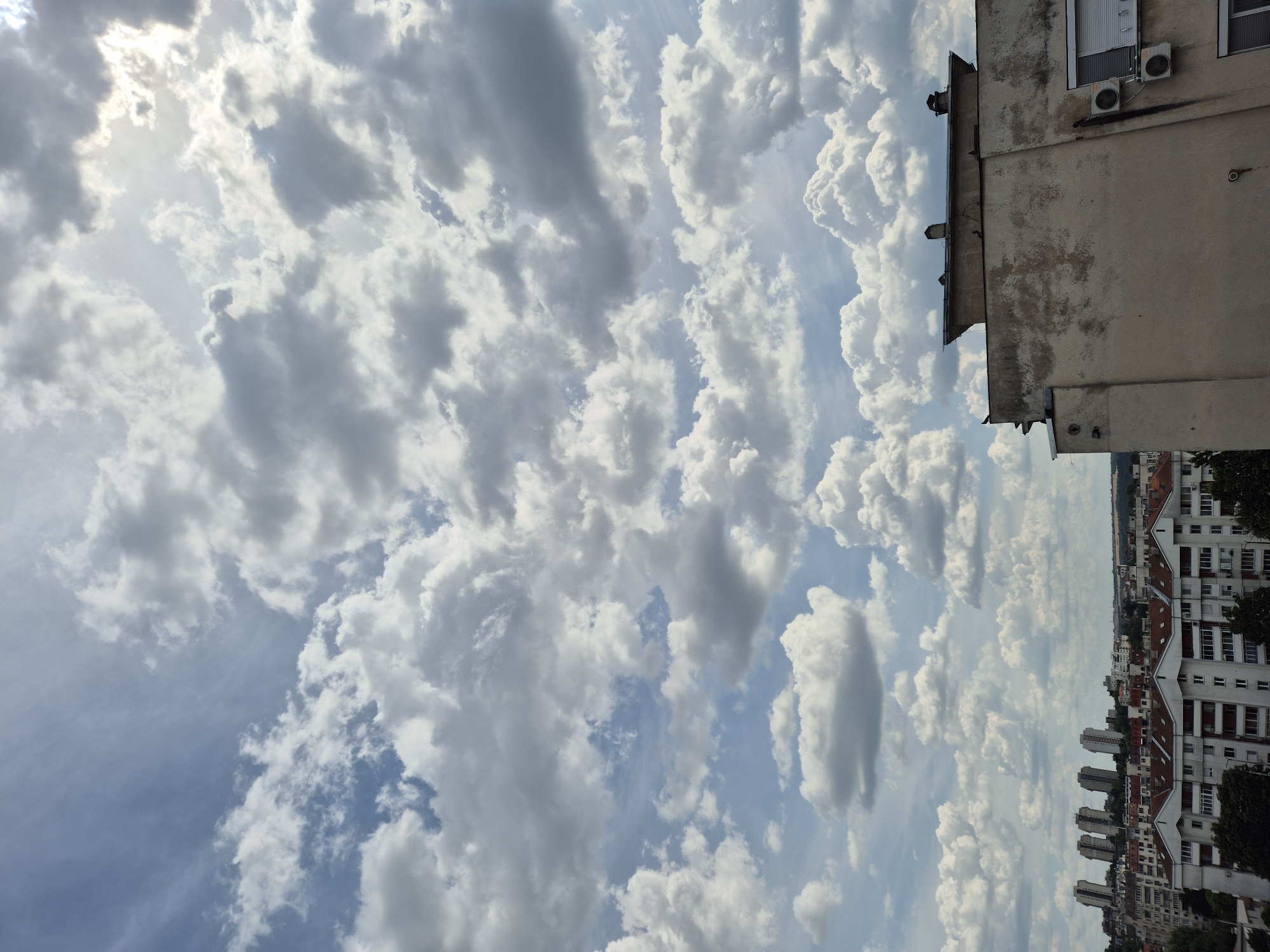The image depicts a city skyline under a cloudy sky. In the foreground, the upper portion of a light-colored building with a weathered facade is visible, featuring two air conditioning units mounted on its exterior wall, along with various rooftop structures including chimneys. Beyond this building, a dense urban landscape unfolds, characterized by a variety of residential buildings with red-tiled roofs and some taller, more modern apartment blocks in the distance. The sky is filled with a mix of white cumulus and gray stratus clouds, with patches of blue sky visible in between. The sun is partially obscured by clouds, creating a bright, diffused light. The scene appears to be a typical urban environment on a day with dynamic cloud formations.