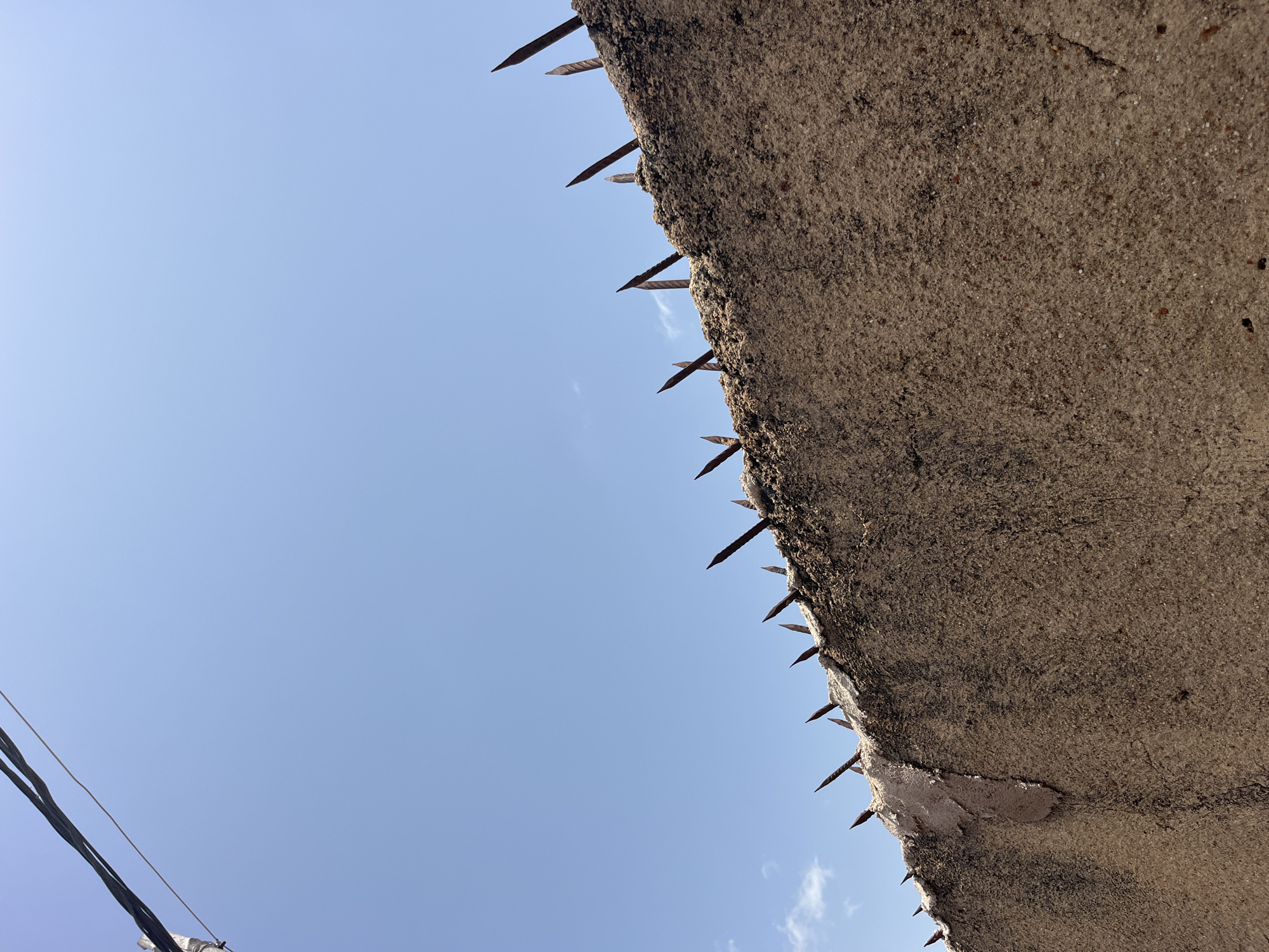 This is a low-angle shot of a concrete barrier with nails protruding from its edge. The barrier occupies the right side of the frame, casting a shadow on the blue sky to its left. The concrete surface is rough and weathered, with some dark stains and cracks. The nails are sharp and rusty, pointing upwards and outwards, suggesting a deterrent or defense mechanism. A few faint clouds are visible in the sky. In the bottom left corner, electrical wires are partially visible, indicating an urban or suburban setting. The lighting suggests it is daytime, likely under a clear sky. There is no text visible in the image.