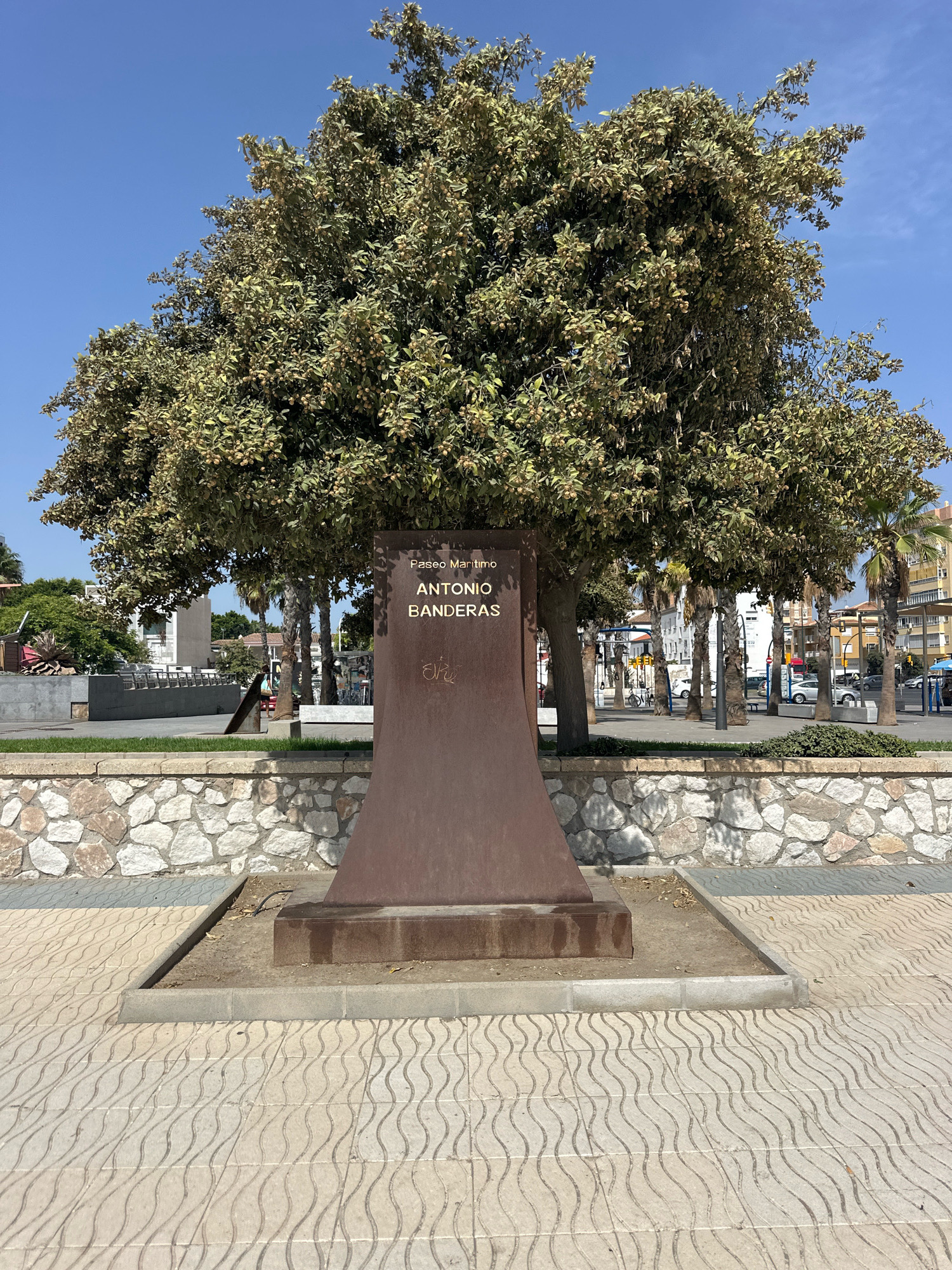 The image shows a monument dedicated to "Paseo Maritimo Antonio Banderas" in the city of Málaga, Spain. The monument is a tall, reddish-brown structure with curved sides, wider at the base and tapering towards the top, inscribed with the name. It is situated in a paved outdoor area with wavy patterned tiles in the foreground and a low stone wall behind it. A large tree with a dense canopy of green leaves and small fruits or nuts stands behind the monument. Palm trees and buildings are visible in the background, suggesting an urban or promenade setting. The sky is clear and blue. There are no people or animals visible in the image.