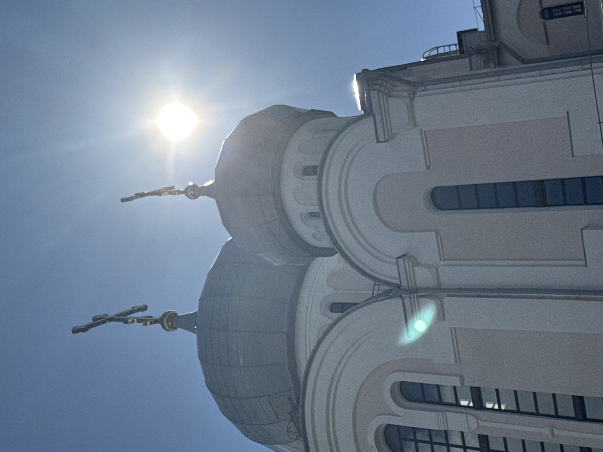 The image depicts the dome and facade of a church, likely an Orthodox Christian church given the presence of crosses atop the domes. The architecture features rounded domes, arched windows, and a pale pink and white color scheme. The sky is clear and bright blue, with the sun prominently visible and casting strong lens flare effects, particularly near the top right of the frame. The sun's rays create a halo effect and illuminate the scene, causing some areas to be overexposed. The image is taken from a low angle, looking up towards the church's domes and the sky. There are no people or other elements of urban life visible in the frame. Without further information, identifying the specific location within Saint Petersburg is not possible based solely on the visual content.