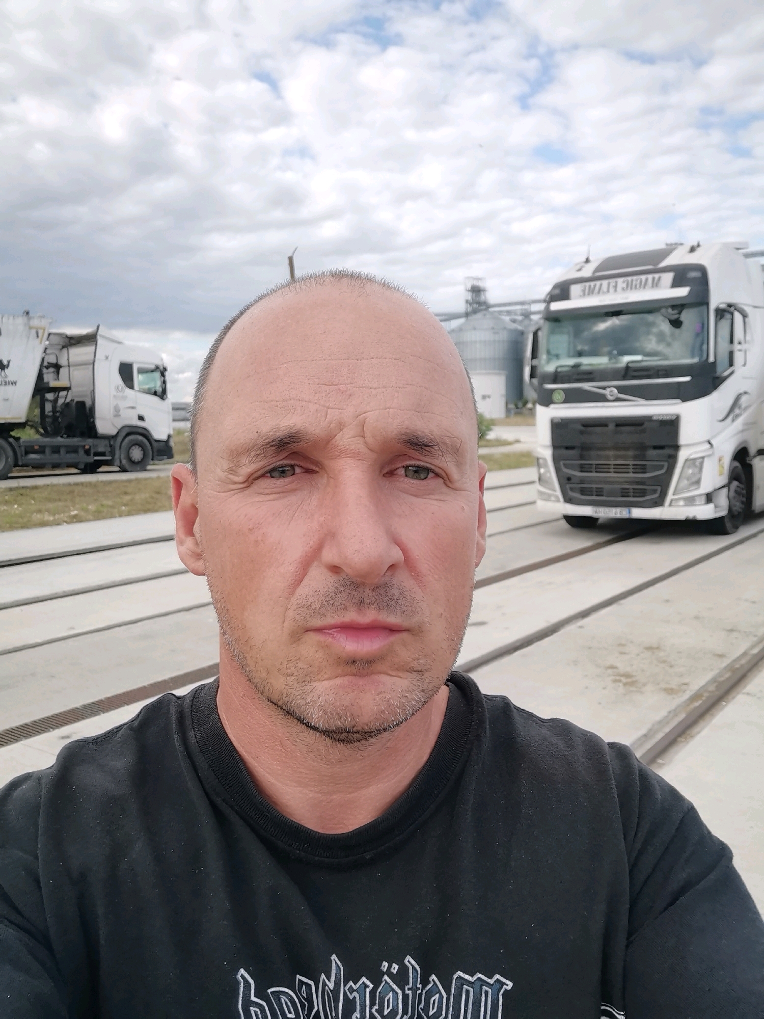 In Aidemir, Bulgaria, a man in a black t-shirt with a band logo is taking a selfie outdoors. He has a shaved head with some stubble on his chin and a serious expression. In the background, two trucks are visible. One is a white garbage truck, and the other is a white Volvo semi-truck with "MAGIC FLAME" written on its windshield. Silo structures are also present in the background, along with a cloudy sky. The man is standing on a concrete surface with parallel metal tracks.