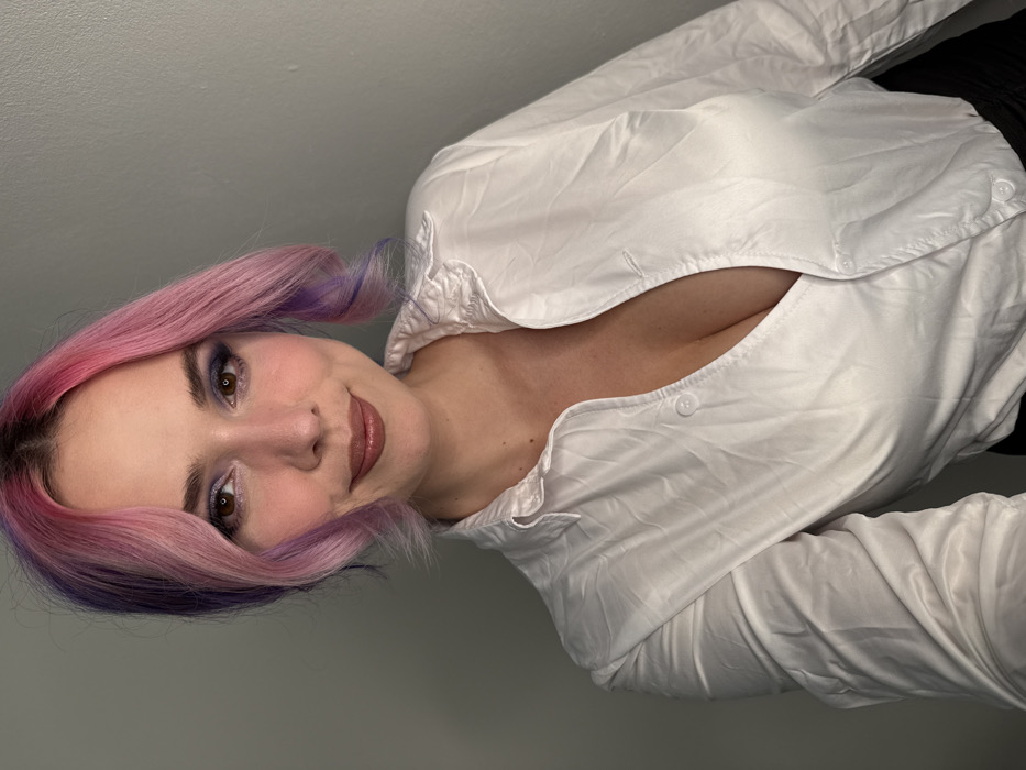 The location of the scene within the City of Zagreb, Croatia, cannot be determined from the visual information provided.
The image depicts a single Caucasian female subject, appearing to be in her mid-20s to early 30s, captured from the chest up. Her medium-length hair is dyed with shades of pink primarily on top and purple underneath, styled with soft waves and parted to her left. Her eyes are brown, accentuated with dark blue and light purple eyeshadow; her lips are coated with a brownish gloss. She is wearing a white, collared shirt, partially unbuttoned to reveal cleavage. A dark garment (possibly a skirt or trousers) is visible at the bottom edge of the frame. The subject gazes directly at the camera with a subtle smile. The background is a plain, light gray wall, indicative of an indoor setting.